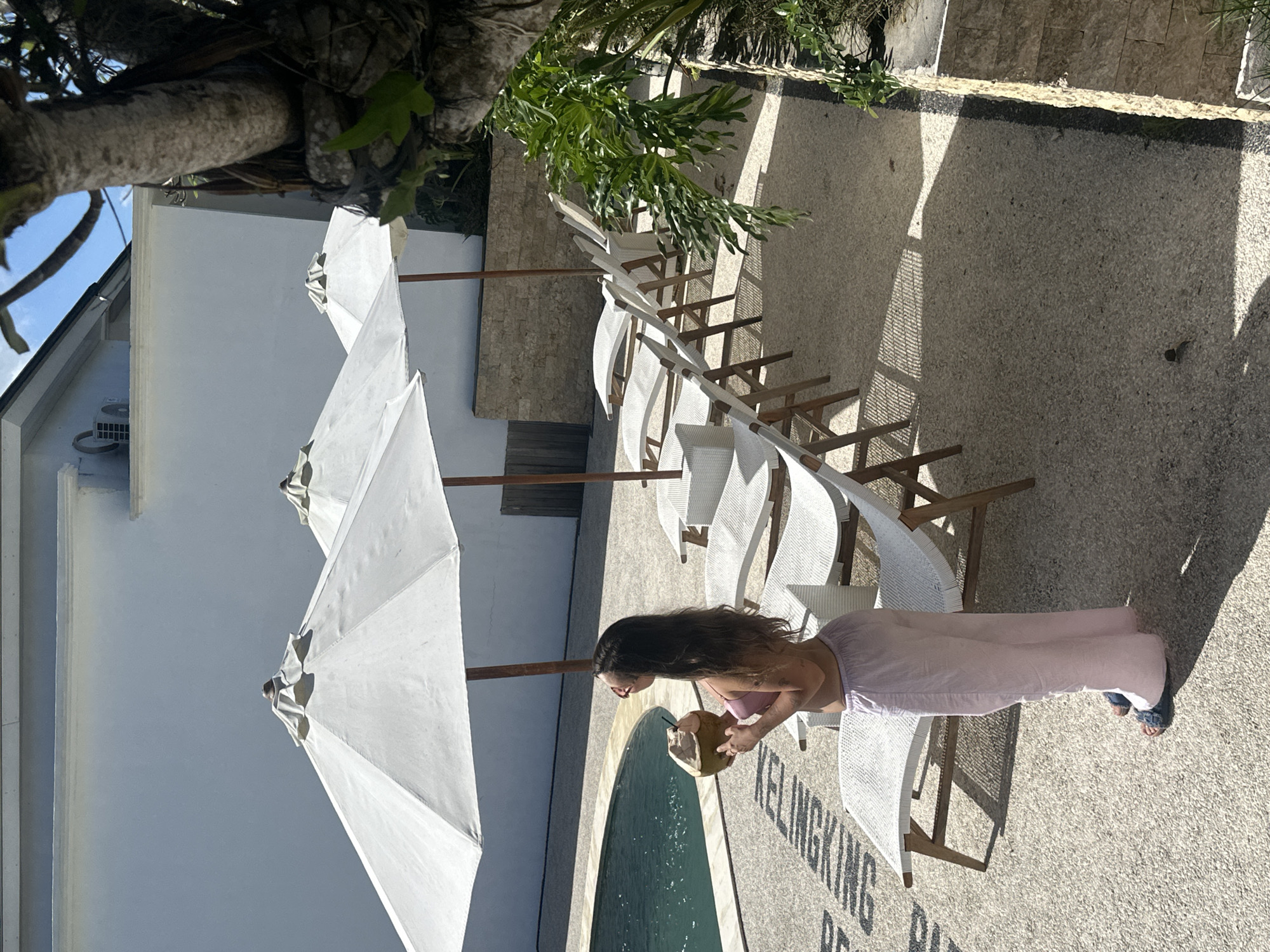 A medium, eye-level shot captures a woman in Nusa Penida, Indonesia, enjoying a coconut drink by a pool. The woman, with long dark hair, wears a light pink top and matching pants. She stands next to a row of white wicker lounge chairs under white umbrellas, positioned along the edge of a curved pool. The pool's edge features the partially visible inscription "KELINGKING RE", indicating the location is likely near Kelingking Beach, a famous site in Nusa Penida. The scene is brightly lit, with harsh shadows cast by the umbrellas and trees. The foreground features the textured ground of the outdoor area, while the background includes a white building with a visible air conditioning unit. A large tree overhangs the top left of the frame.