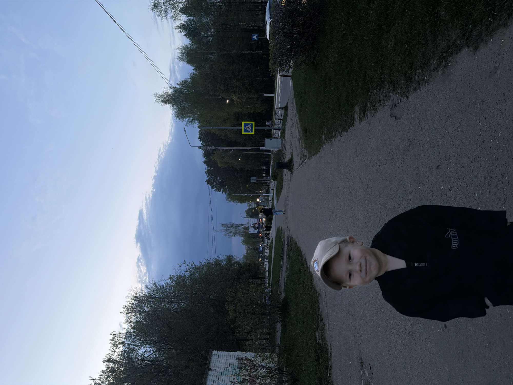 A photograph taken in Chaikovsky, Russia shows a young boy in a black Puma jacket and light colored baseball cap standing on a paved path. A street, crosswalk, and trees are visible behind the boy. Streetlights line the street and some buildings are visible in the distance. The sky is light blue and slightly cloudy.