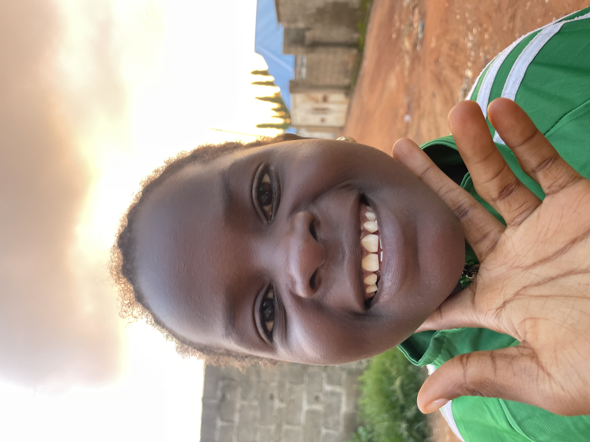 The media file features a close-up, rotated portrait of a young girl in Rido, Nigeria. She has dark skin and is smiling broadly, showing her teeth. Her left hand is raised and positioned near her face. She is wearing a green shirt with white stripes on the collar. The background suggests an outdoor setting with a dirt path, brickwork, and buildings under a bright, hazy sky, possibly indicating late afternoon or early morning due to the warm light. The overall mood is cheerful and lively. There is no visible text in the image.