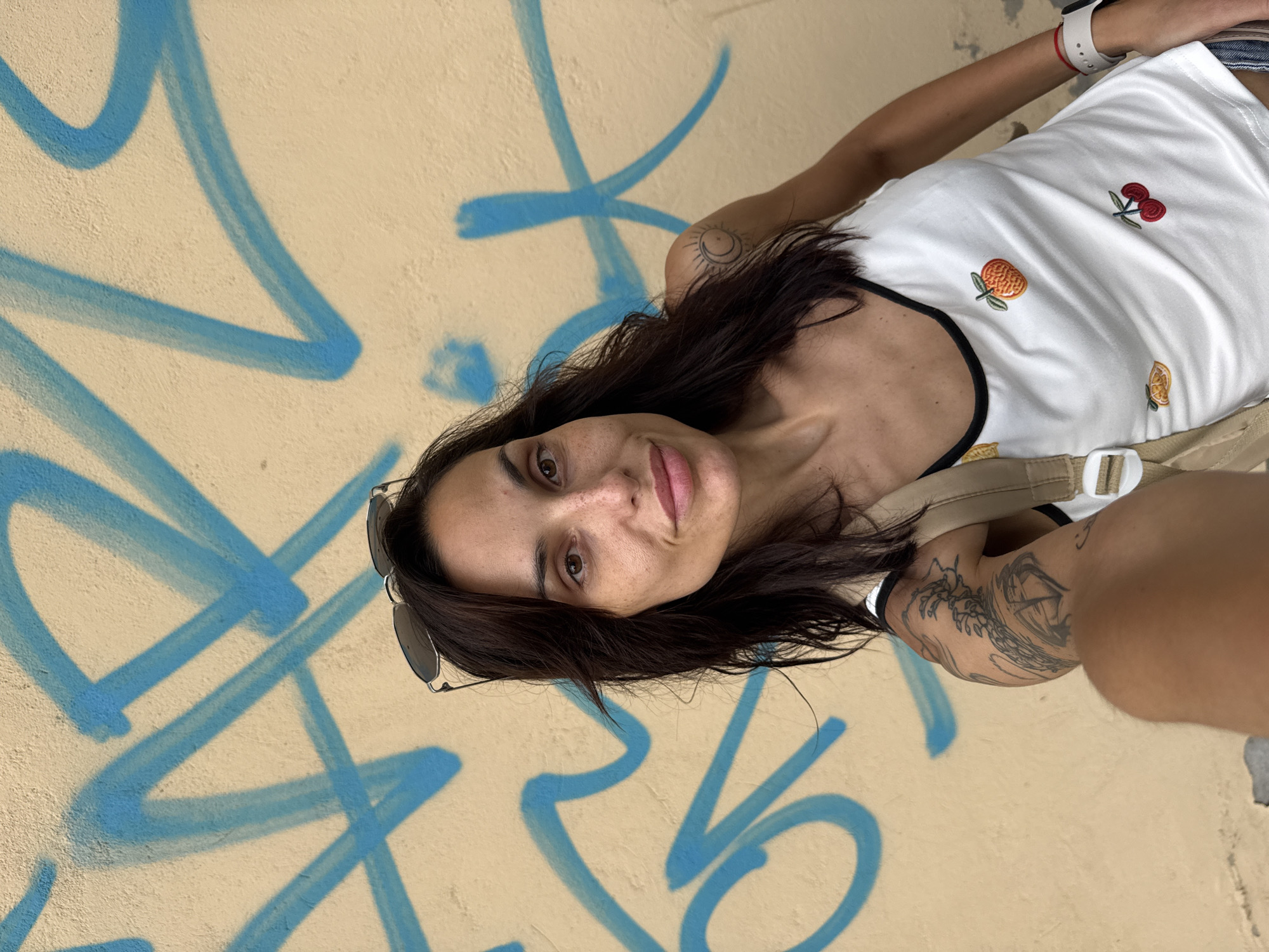 An adult female is captured in a selfie-style photograph against a light beige wall covered in blue graffiti. She has dark hair, dark eyes, and freckles, with sunglasses resting on her head. She is wearing a white tank top with black trim, embroidered with an orange and cherries, and a light-colored backpack strap is visible over her right shoulder. A sun and crescent moon tattoo is on her right upper arm, and a more intricate tattoo is visible on her left upper arm. A white smartwatch and a red string bracelet are on her left wrist. The image appears to be taken outdoors in the city of Pleven, Bulgaria.