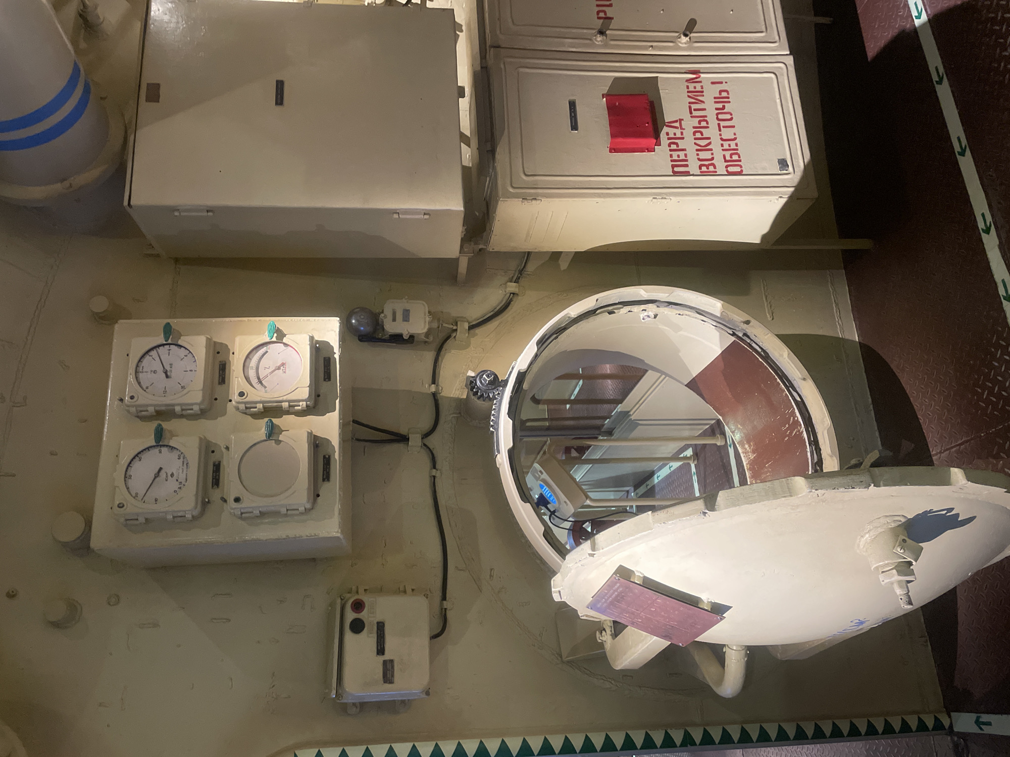 The image captures an interior view of what appears to be a submarine hatchway or a similar compartment within a museum or historical exhibit in Kronstadt, Russia. The scene is dominated by industrial elements and control panels.
In the foreground, an open, circular hatch door, painted a light cream color, is swung outwards to the left. It has a red panel attached to its inner surface. The hatchway itself is a large, ring-shaped opening with a reddish-brown interior lining. Beyond the hatch, a dimly lit corridor or passage can be seen, with a railing and what appears to be a metal staircase or ramp. Green directional arrows are painted on the floor, indicating pathways.
On the wall to the left of the hatch, a cream-colored electrical junction box with two black circular openings is mounted. Above this, a panel holds four circular gauges. Two of these gauges are visible and appear to be pressure or temperature indicators, with needles pointing to readings. Below these gauges are two square panels, also cream-colored.
To the right of the gauges, a large, tall, cream-colored cabinet or control panel unit is mounted on the wall. Below this, another set of cream-colored cabinets are stacked, with red lettering on the front of the lower cabinet that reads "ПЕРЕД ВСКРЫТИЕМ ОБЕСТОЧЬ!", which translates to "Before opening, de-energize!". A small red box is mounted above this lettering.
The walls and ceiling are painted a muted, utilitarian color, possibly a light olive or beige. Metal pipes, one with blue and white stripes, are visible towards the top of the frame. The lighting is somewhat dim, casting shadows that emphasize the metallic and industrial textures of the environment. No people are present in the image. The overall impression is one of a functional, historical, and possibly preserved technical space.