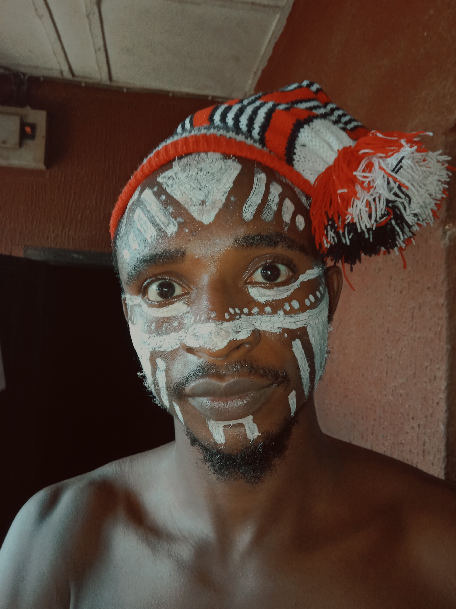 A close-up shot depicts a man wearing a red, black, and white knit hat with pom-poms. His face is painted with white markings. He has dark skin, a beard, and a mustache. The background consists of a red-painted wall and part of a white ceiling. Location is in the city of Unknown, Unknown.