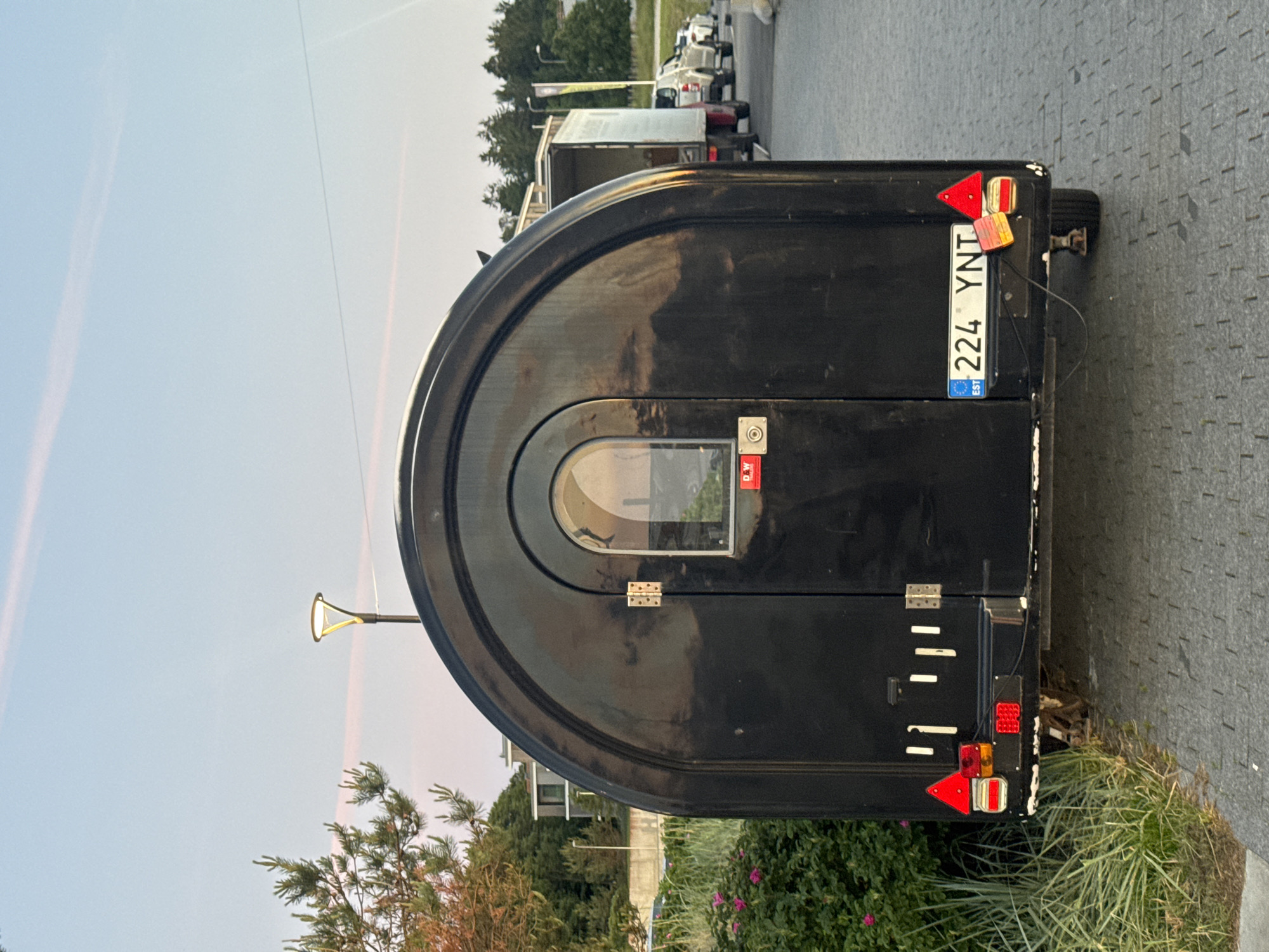 A black trailer with an arched roof and a rounded door is parked on a paved area, likely in Tallinn, Estonia, as indicated by the license plate "224 YNT EST". The trailer has red and amber reflectors and a white license plate. Behind the trailer, a white box truck is visible, followed by other vehicles. Green trees and a building can be seen in the background, and the sky is a pale blue with wisps of pink clouds. The lighting suggests it is late afternoon or early evening.