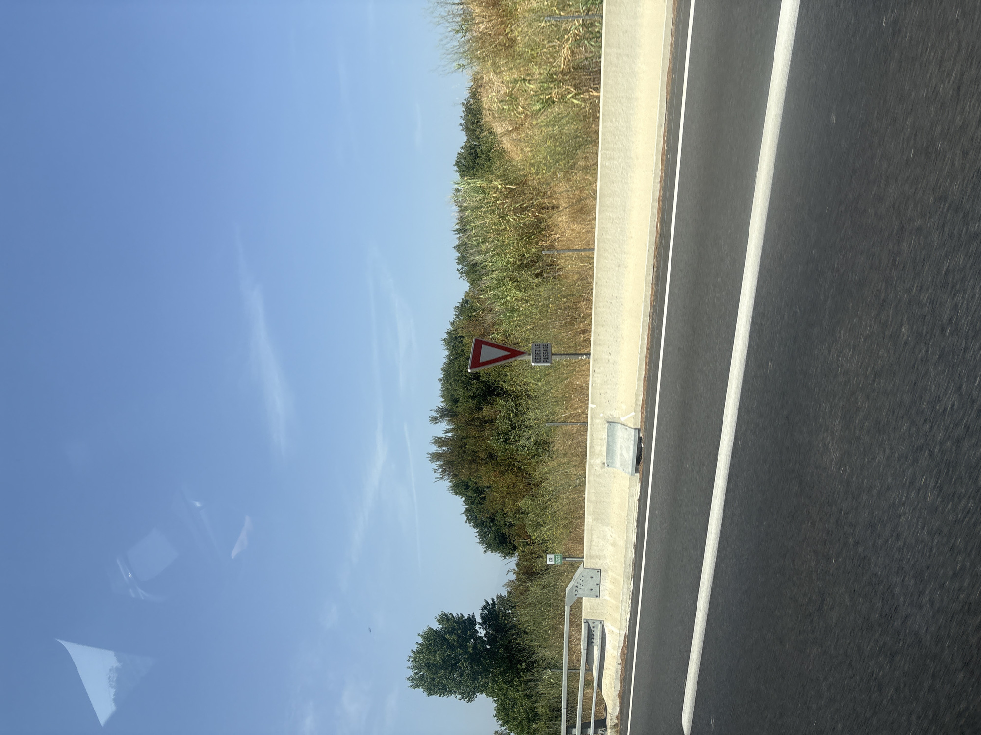 The image depicts a view from a moving vehicle on a highway, likely in Pia, France, given the presence of a French "cédez le passage" (yield) sign. The foreground is dominated by the dark asphalt of the road, marked with a white line. A low, light-colored concrete barrier runs along the side of the road, separating it from the natural environment.
Beyond the barrier, the landscape is characterized by dense vegetation, including trees and tall, dry grasses. The sky is a clear, bright blue with a few wispy clouds. A triangular red and white "yield" sign with French text is prominently visible, indicating an upcoming intersection or merging point. A small green sign with white text and symbols is also present on a post near the barrier. The overall scene suggests a sunny day in a rural or suburban area.