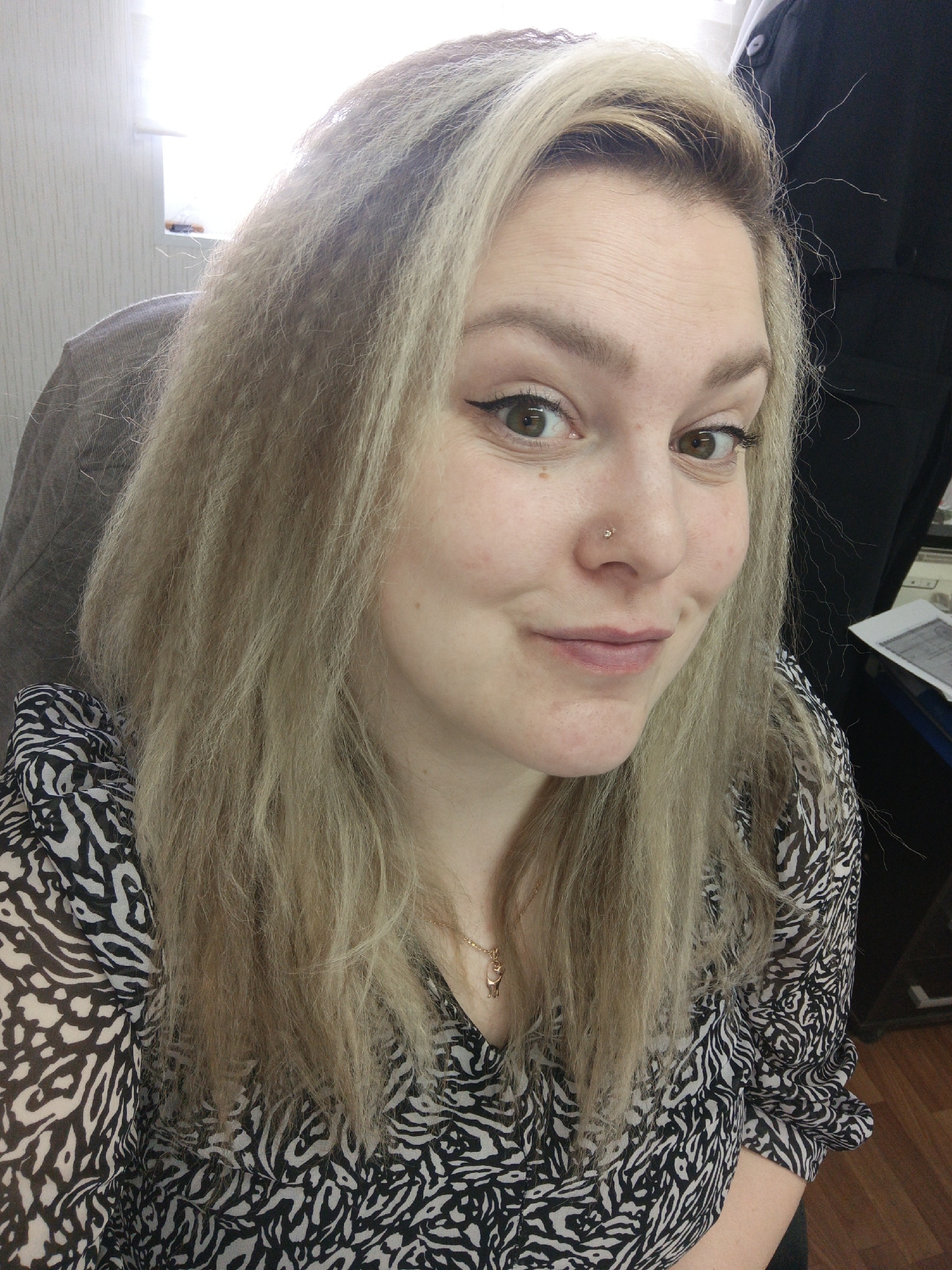 A woman with light blonde, crimped hair is in a room. She is wearing a black and white patterned blouse with a v-neck. She has a nose piercing and is wearing eyeliner. A window is visible in the background. A portion of a dark-colored garment is visible on the right side of the frame. A chair can be seen on the left. There is no definitive evidence in the image to confirm the location as the city of Noviy, Russia.