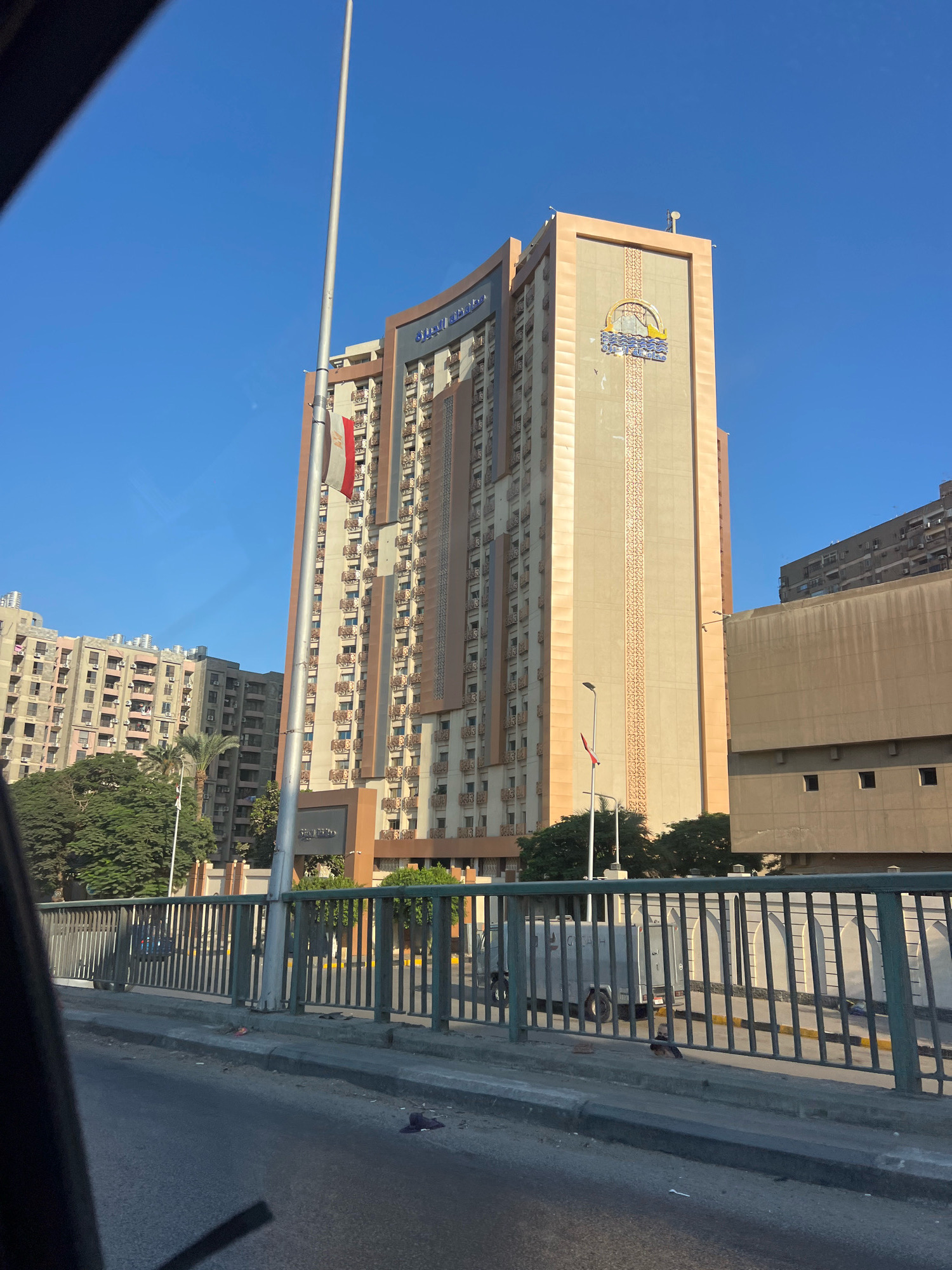 The photograph captures a view of a tall, modern building in Giza, Egypt, on a clear, sunny day. The building, likely a hotel or office complex, has a beige facade with decorative vertical patterns and a curved upper section. An Egyptian flag flies from a flagpole in front of the building. In the foreground, a metal railing separates a sidewalk from a road, suggesting the photo was taken from a moving vehicle or a bridge. Adjacent to the main building are other multi-story residential or commercial structures, as well as trees, indicating an urban environment. A white armored vehicle is visible behind the railing. The overall scene depicts a section of Giza's cityscape with architectural elements characteristic of the region.