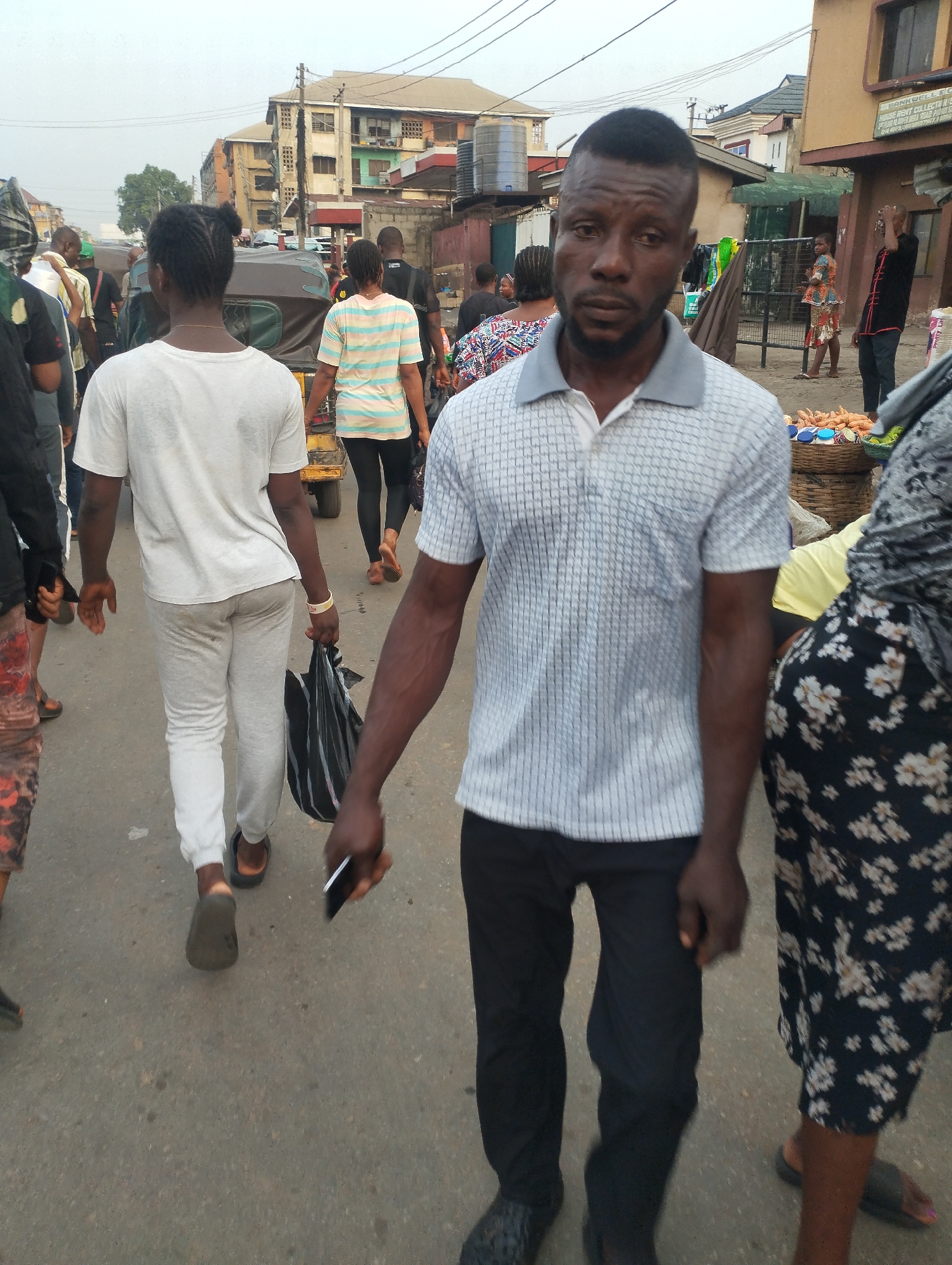 The image depicts a street scene in Fegge, Nigeria. A man wearing a light blue and white striped polo shirt and black pants is walking towards the camera, holding a black smartphone in his right hand. To his left, a woman in a white shirt and light gray pants walks away from the camera, carrying a black plastic bag. Other people are walking in both directions along the street, including a woman in a striped shirt and a woman in a floral print dress. A yellow tricycle is also visible in the background. A building can be seen in the background, and vendors can be seen.