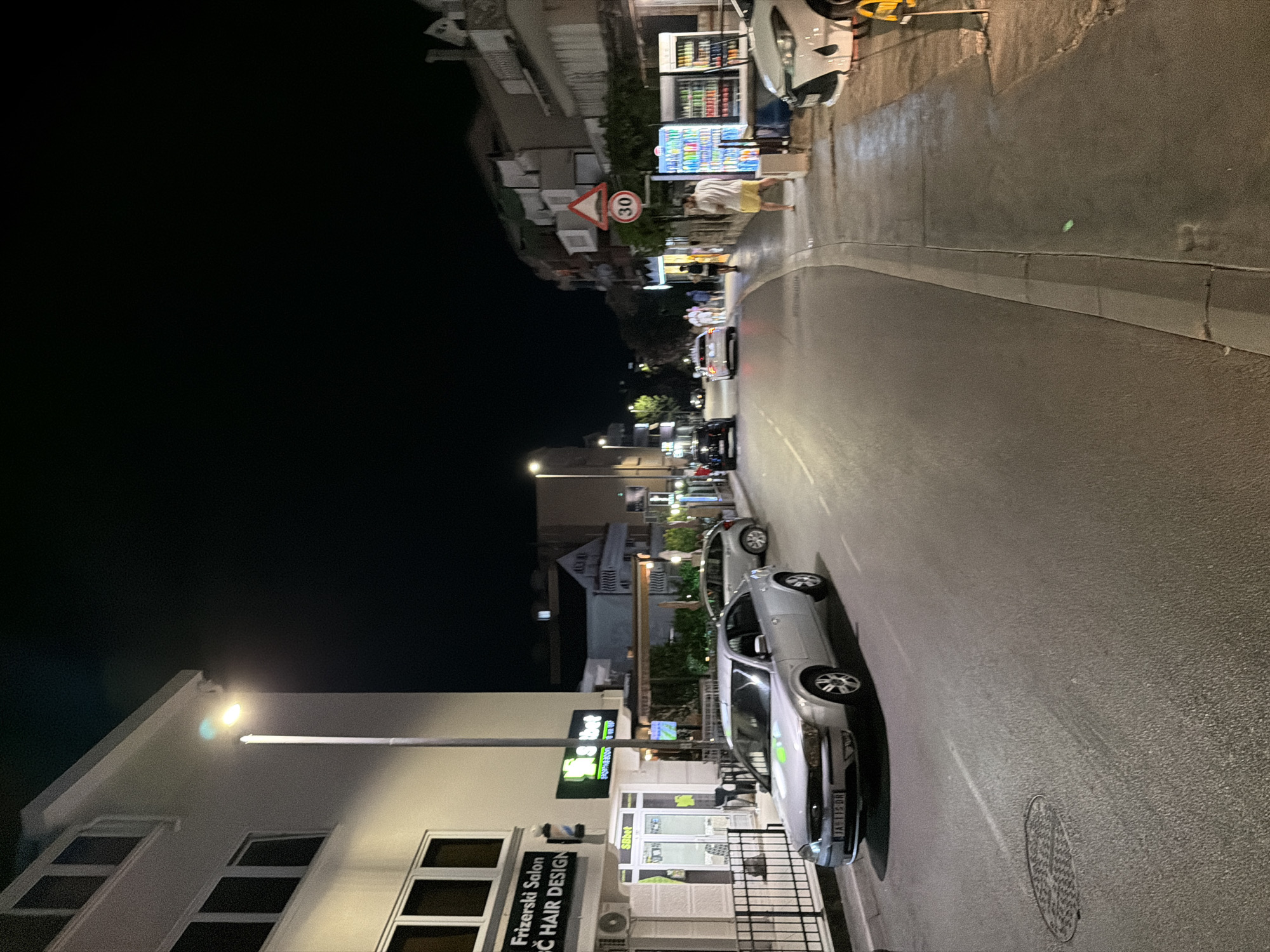 The image shows a street scene at night in Budva, Montenegro. The street is paved and curves to the right. On the left side of the street, there is a multi-story building with windows. A street lamp illuminates the area. Below the building, there are storefronts, including a "Frizerski Salon" (hair salon) and a "Sbbet" establishment. Several cars are parked or driving on the street. In the foreground, a silver sedan is visible. On the right side of the street, there is a sidewalk with several people walking and standing. There are also vending machines stocked with drinks. Buildings line the right side of the street. A speed limit sign indicating "30" is mounted on a pole. The overall atmosphere is that of a busy commercial street at night.