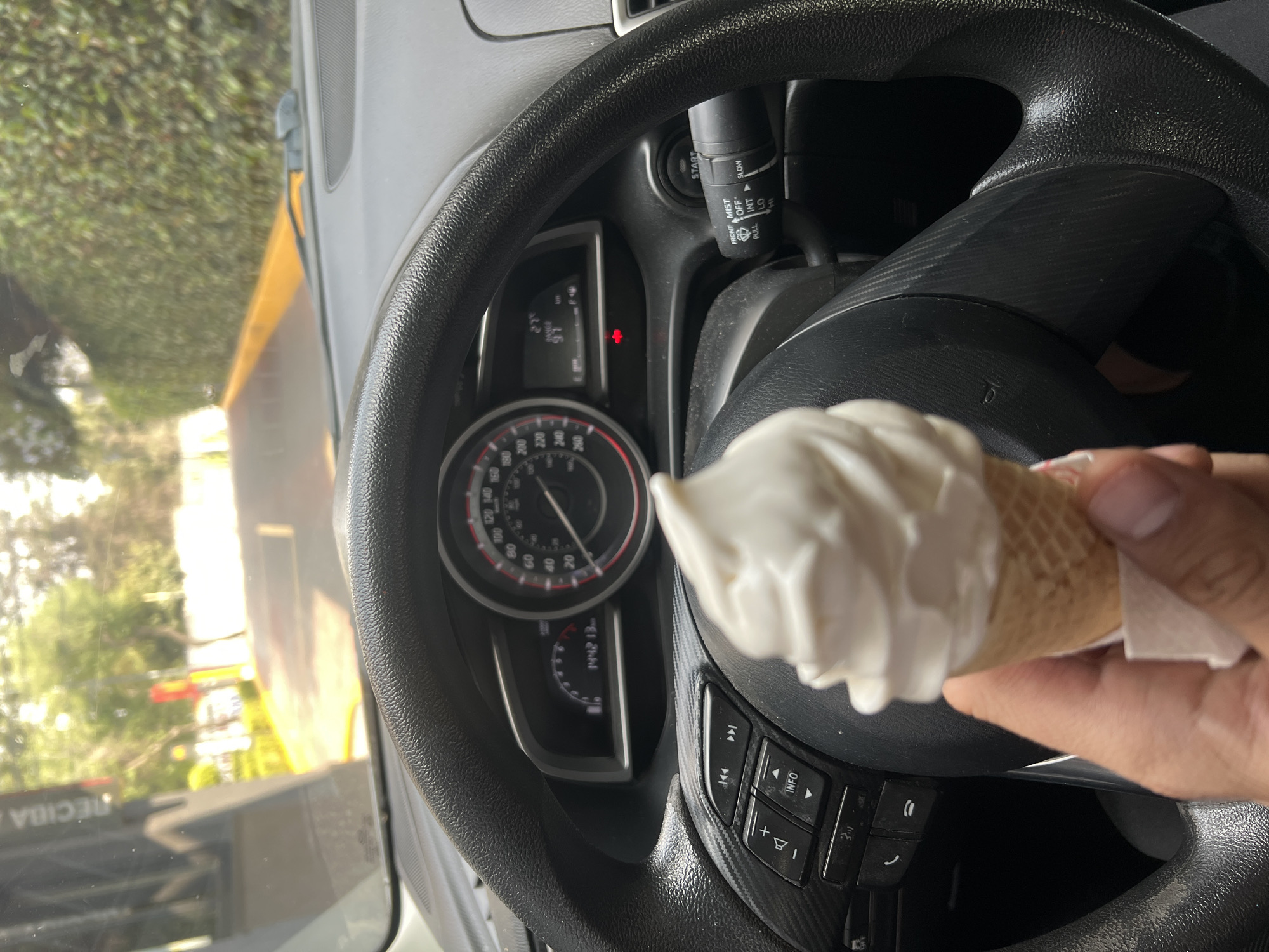 A person sits inside a vehicle, holding an ice cream cone in their right hand in front of the steering wheel. The dashboard and instrument panel are visible. Outside the window, foliage and buildings can be seen. The windshield reflects other vehicles on the road. The OCR provides the city name "RECIBE" as part of the signage in Mexico City.