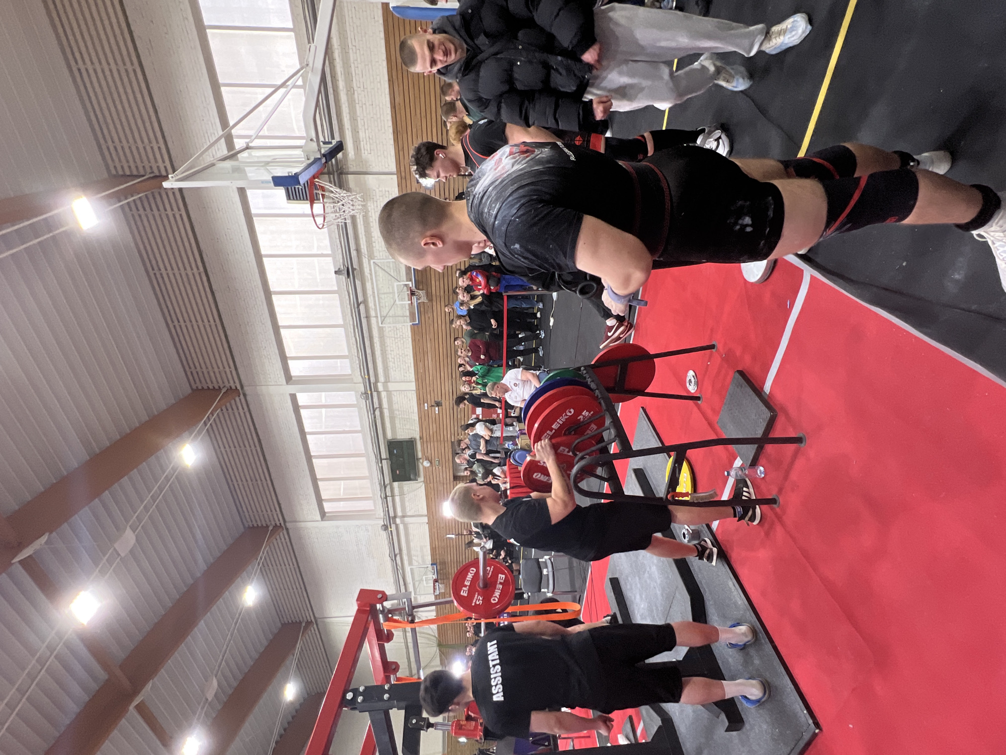 The image depicts a weightlifting competition held indoors, potentially within a gymnasium, in Saint Petersburg, Russia. The scene showcases a weightlifter preparing to lift a loaded barbell, with multiple weight plates visible. Two assistants are present, likely assisting with the loading and spotting. Spectators are observing the competition from the background. The environment includes a basketball hoop, typical of a gymnasium setting. The flooring consists of a red platform with black mats, demarcating the weightlifting area.