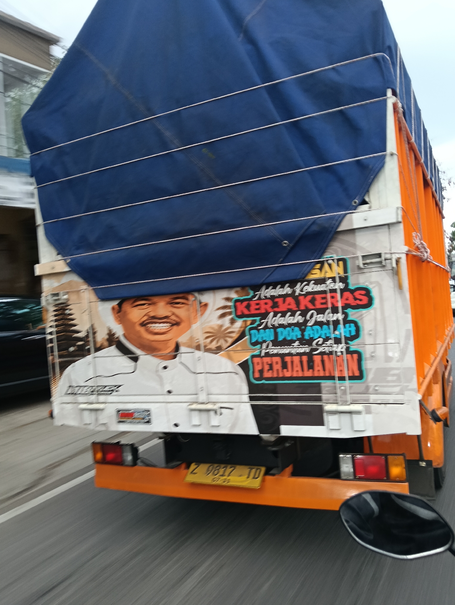 A medium shot captures the rear of an orange truck with a blue tarp cover, its side adorned with a graphic of a smiling man in a white shirt and a black collar. The man's portrait is superimposed on a backdrop of a pagoda-like structure and palm trees. To the right of the portrait, text in Indonesian reads, "Adalah Kekuatan KERJA KERAS Adalah Jalan DAN DOA ADALAH Penentu Satuan PERJALANAN." (Translation: "Is the strength of HARD WORK Is the way AND PRAYER Is the determinant of the JOURNEY.") The truck's rear bumper is orange, and a yellow license plate with the characters "Z 9817 TD" is visible. The scene appears to be on a road, with a blurred view of the surrounding environment indicating motion. The overall lighting suggests daytime. The location is the city of Cintaraja, Indonesia.