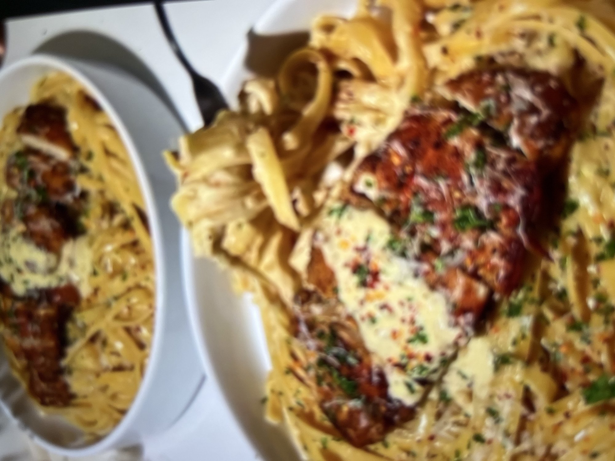 The image shows two bowls of pasta with what appears to be chicken on top. The pasta is a fettuccine or linguine style and is coated in a creamy sauce. There are flecks of green herbs and red pepper flakes visible on the pasta and the chicken. One bowl is in the foreground and is in sharp focus, while the other bowl is in the background and is slightly blurred. The setting appears to be a restaurant. There is no information available to suggest this image is located in Abuja, Nigeria.