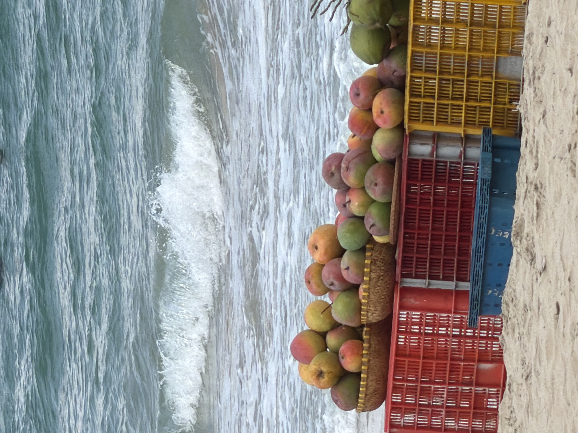 Seashore scene in Cam Lâm, Vietnam. A stack of crates containing fruit, including mangoes and coconuts, is arranged on a sandy beach near the water's edge. The crates are yellow, red, and blue. Waves wash onto the shore.