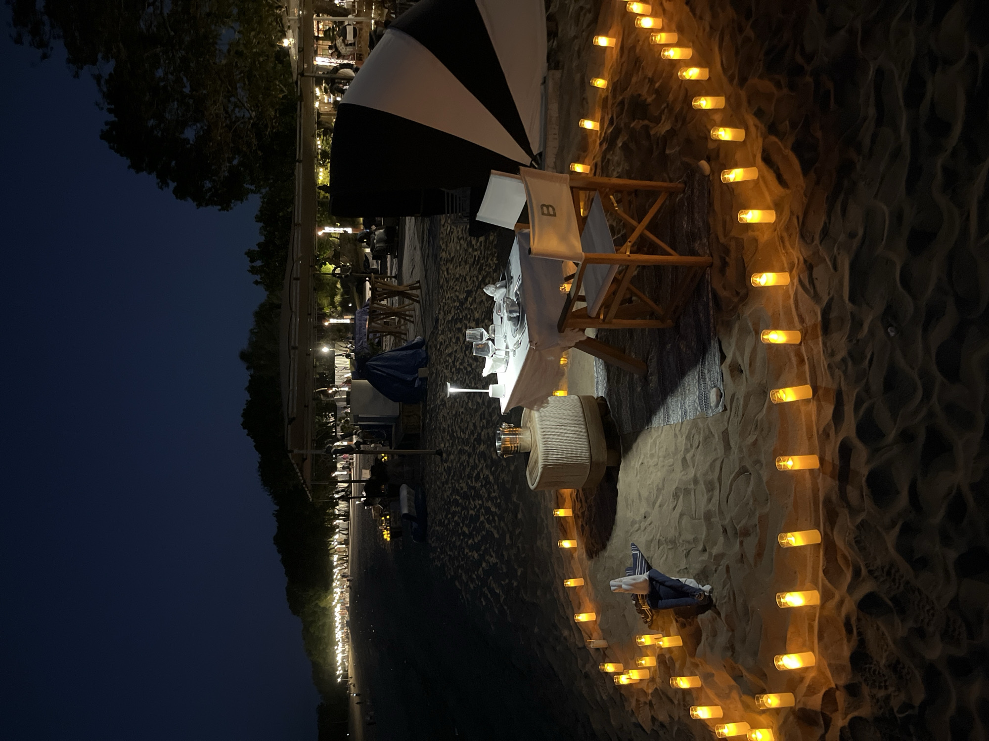 A romantic beach dinner is set up on the sandy shore of Kuta Selatan, Indonesia at night. The scene is illuminated by dozens of glowing candles arranged in a circular pattern around a small dining area. In the center of this circle, a white-clothed table is set for two with plates, glasses, and napkins. A director's chair with the initial "B" on it is placed at the table, facing the ocean. To the left of the table, a small stool holds an ice bucket and a bottle. In the background, a row of trees and structures line the beach, with more lights visible in the distance. A large black and white striped umbrella is visible behind the chair, and a towel and a small rolled-up mat lie on the sand to the left of the table. The night sky is dark and clear.