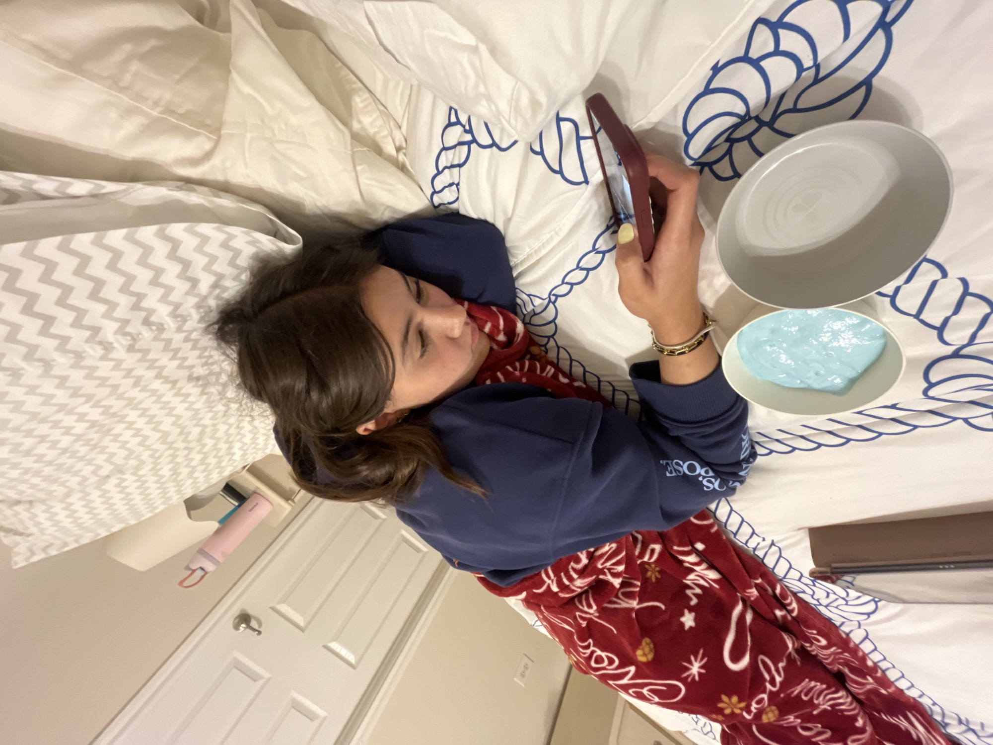 A medium shot shows a woman lying on her back on a bed, using a mobile phone. The bedding consists of white sheets with a blue rope-like pattern, a pillow with a gray chevron pattern, and a red blanket with white writing and designs. To her right are two bowls, one empty and gray, the other containing a light blue substance. The room's wall is light beige, and a white door and a pink water bottle are partially visible in the background. The scene is likely inside a residence, but a precise location within San Antonio cannot be determined from the image.