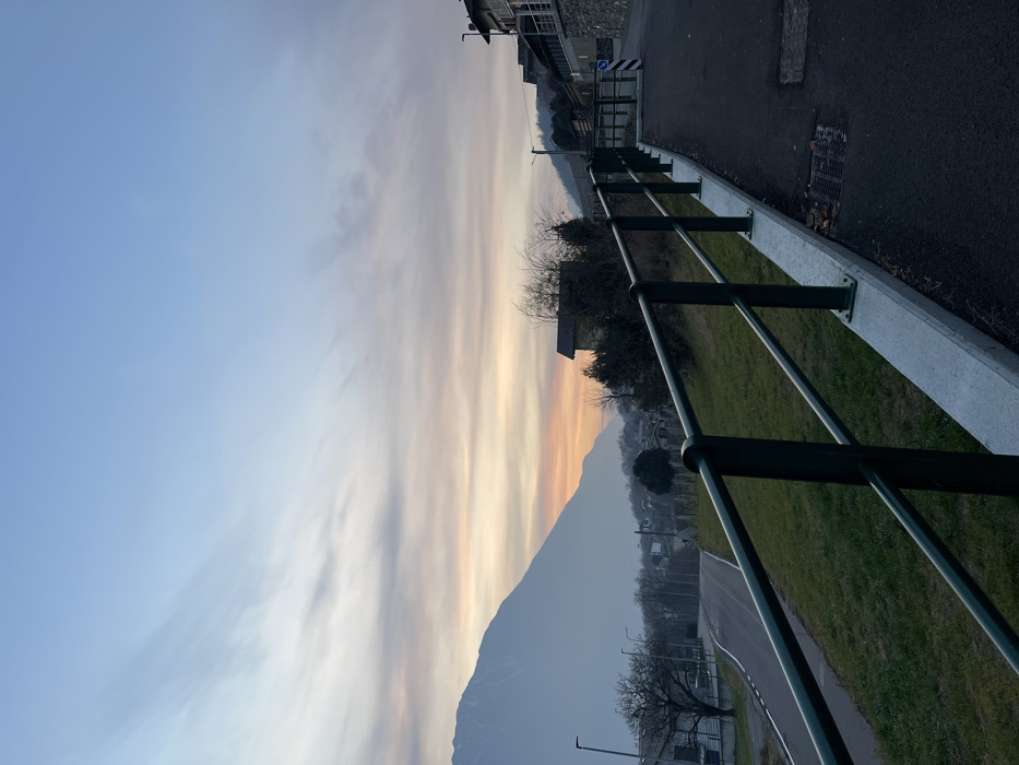 This image depicts a serene landscape scene, likely at either sunrise or sunset, over Castione, Italy. The sky dominates the upper half of the frame, displaying a beautiful gradient from light blue at the top to warm orange, pink, and yellow hues near the horizon, characteristic of twilight. Long, wispy clouds are stretched across the sky, catching the soft, warm light.
A prominent mountain range forms a dark silhouette against the vibrant sky, creating a strong horizon line. At the foot of these mountains, structures of a village or town are visible, nestled into the landscape.
In the foreground on the right, a dark green metal railing with regularly spaced posts acts as a barrier, running alongside a paved road or path. The railing is anchored into a light-colored concrete curb. The asphalt path features faint white road markings and a visible drainage grate. Beyond the railing, a gentle slope of green grass extends towards the mid-ground. Scattered, bare-branched trees are seen on the grassy slope and in the village area, suggesting a winter or early spring setting. To the far right, a building with a stone facade and a dark roof is partially visible.
No people are present, and no specific activities are taking place, contributing to a tranquil and still atmosphere. A small blue and white chevron sign can be seen near the road in the mid-ground. The overall impression is one of peaceful natural beauty transitioning between day and night.