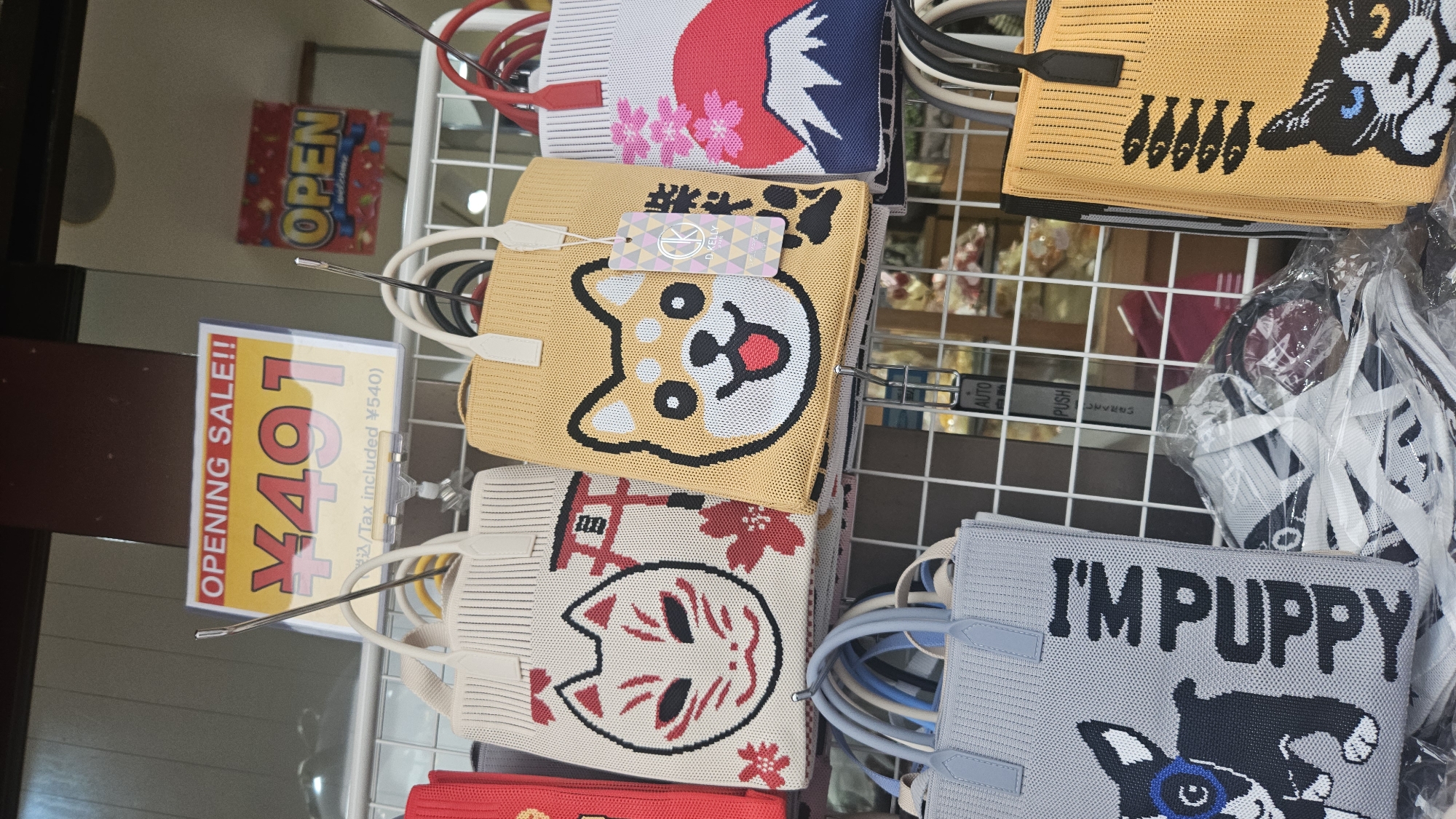 A retail display features knitted tote bags with graphic designs of animals and Japanese cultural motifs. In the upper left corner, a sign announces "OPENING SALE!!" with a price of "¥491 (Tax included ¥540)". To the right, a colorful "OPEN" sign is visible. The bags are displayed on a tiered metal rack.
The bags shown include:
- A white bag with a red fox mask design and a red torii gate symbol.
- A yellow bag with a Shiba Inu dog face.
- A red bag with white trim.
- A blue-grey bag with a black and white dog design and the text "I'M PUPPY".
- A yellow bag with a black and white cat face.
- A white bag with a red Mount Fuji and cherry blossom design.
The bags are displayed in what appears to be a shop or market stall, potentially in Kamakura, Japan, given the Japanese text and motifs on some of the items, though the specific location within Kamakura is not determinable from the image alone. The overall scene suggests a commercial environment with a focus on novelty or character-themed merchandise. There are no people clearly visible in the foreground or interacting with the merchandise. The lighting suggests an indoor setting with artificial light.