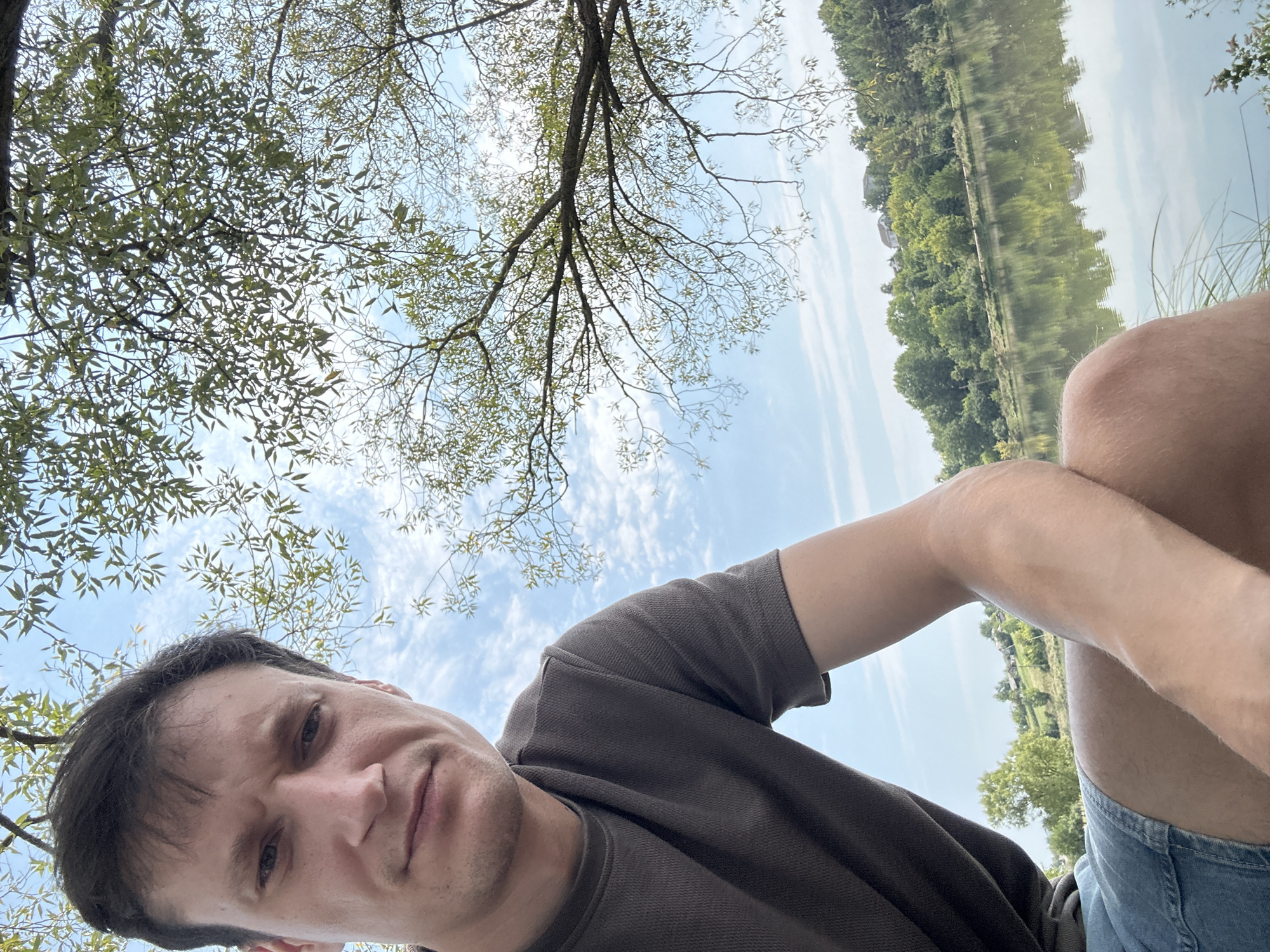 A close-up, selfie-style photograph shows a man from the chest up, positioned outdoors. He has short, dark brown hair and fair skin, with a neutral expression. He is wearing a dark grey, waffle-knit t-shirt and denim shorts.
The background reveals a tranquil scene beside a body of water, likely a lake or river, reflecting the sky and surrounding landscape. On the far bank, a tree-lined hillside is dotted with several houses. The sky is partly cloudy, with streaks of white clouds against a pale blue backdrop. The branches of a leafy tree hang down from the top of the frame, partially obscuring the sky.
The photograph appears to have been taken on a clear, sunny day in the village of Makiv, Ukraine, as indicated by the natural, rural setting with a prominent water feature and residential structures on the distant hills. The overall ambiance is one of relaxation and enjoyment of nature.