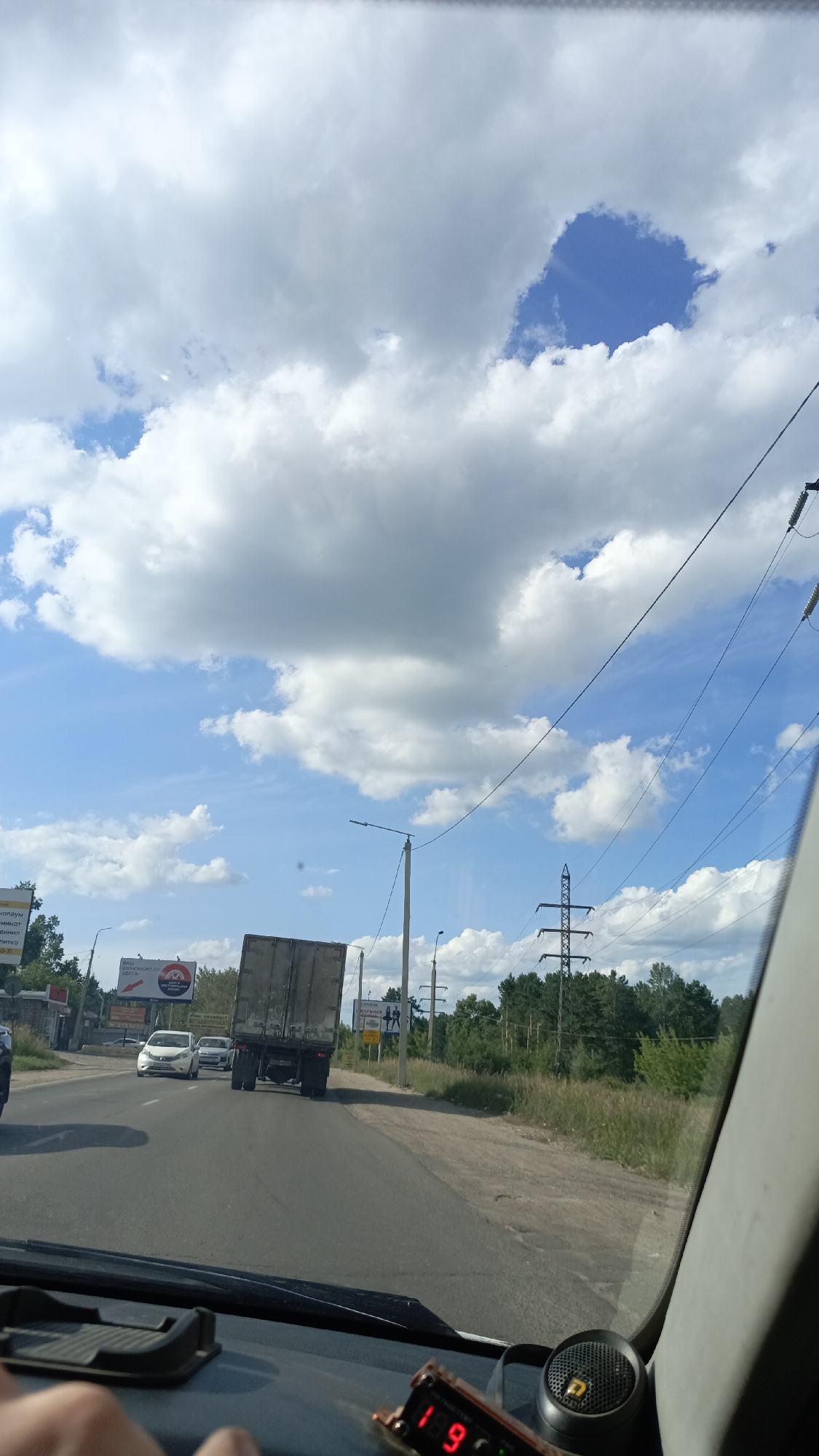 The image depicts a view from inside a vehicle, looking forward and slightly upward. The sky is predominantly blue with large, white, fluffy clouds. A road with two lanes is visible, with a truck in the right lane ahead. On the left side of the road, there are some buildings and billboards, including one with Russian text and images. Trees line the right side of the road, and power lines and poles are visible in the distance. The dashboard of the vehicle is partially visible in the foreground, with a digital display showing "18.9". This image was taken in the city of Angarsk, Russia.