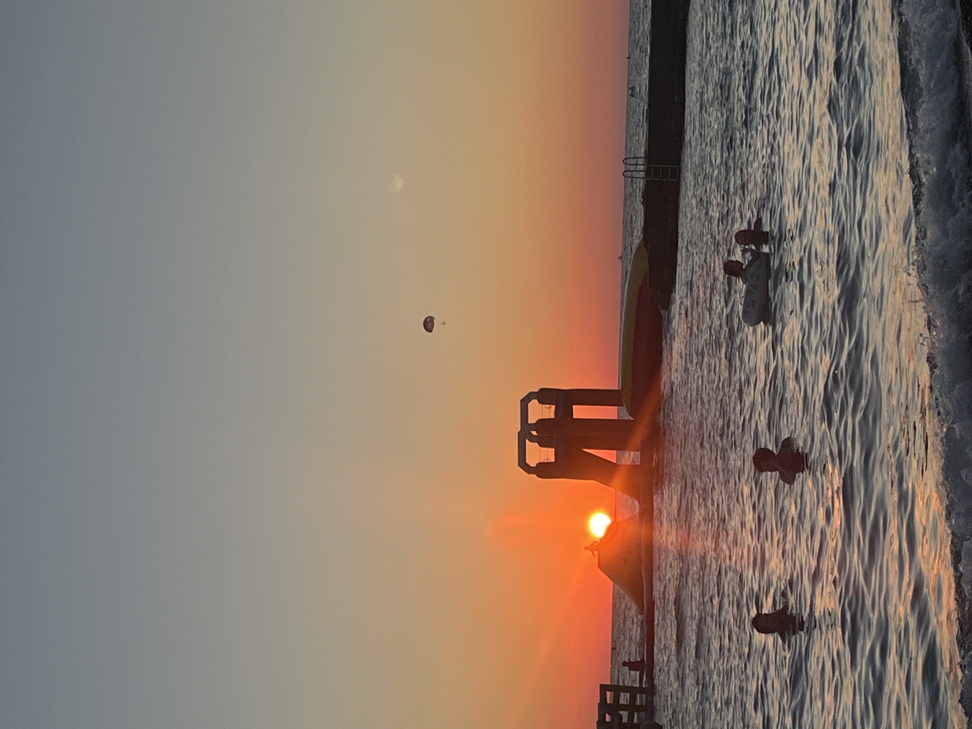 The image depicts a sunset at a beach resort in Sochi, Russia. The sun is setting over the Black Sea, casting a warm orange glow across the sky. In the foreground, several people are swimming in the water, some with inflatable toys. In the middle ground, there is an inflatable water slide and a platform structure. In the distance, a parasailer is visible in the sky, and a few boats are out on the water. The overall scene conveys a sense of relaxation and leisure on a summer evening.
