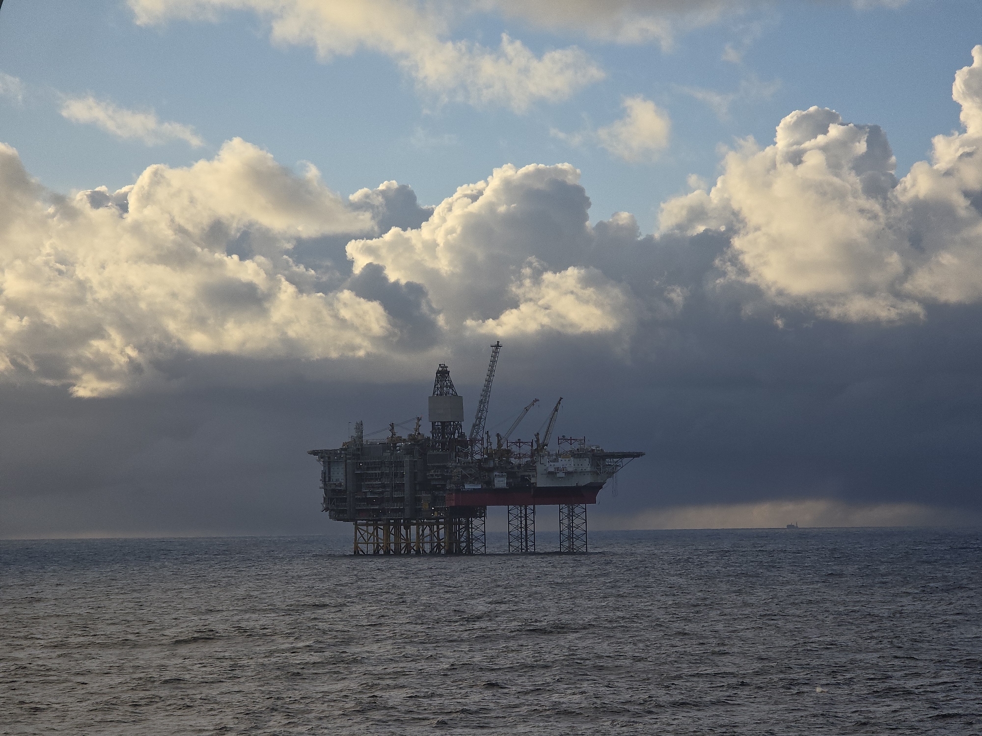 The image shows an offshore oil rig in the ocean under a cloudy sky. The rig is a complex structure with multiple levels, cranes, and a derrick. It is supported by yellow and gray legs that extend down to the seabed. The ocean is choppy with visible waves. In the distance, a small vessel is visible on the horizon. The clouds are a mix of dark and light gray, with some patches of blue sky visible. There is no indication of the city or location.
