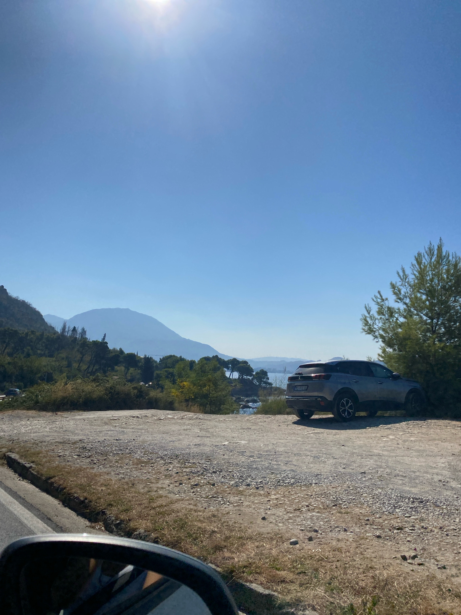 The image depicts a sun-drenched day in Sutomore, Montenegro. A silver SUV is parked on a gravel lot overlooking a body of water and distant mountains. The car is positioned to the right of the frame, partially obscured by a tall, leafy tree. In the foreground, a dirt road with sparse vegetation lines the bottom of the image, with a car's side mirror visible on the left. The sky is a clear, bright blue, with the sun prominently positioned in the upper left quadrant. The landscape features rolling hills covered in trees and shrubbery, with the silhouette of mountains in the background, suggesting a coastal or mountainous region. There are no people visible in the image.
