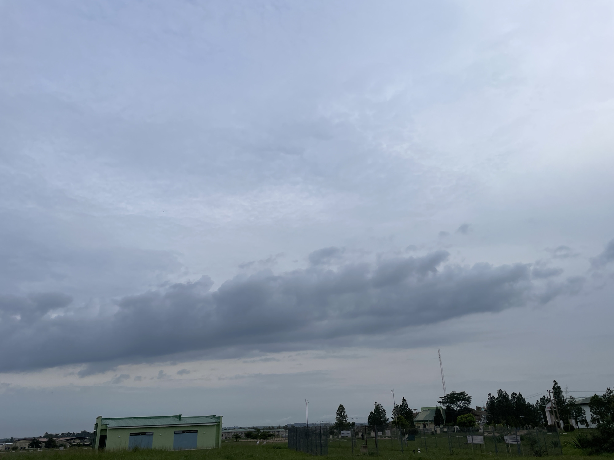 The image depicts an overcast sky over a grassy field in Durumi, Nigeria. In the foreground, a small, light green building with a dark green roof is visible on the left. The building has two light blue doors. Further back, a line of trees and several other buildings are scattered across the landscape. A tall, thin radio tower stands out among the trees. The ground is covered in green grass, with some fencing and poles visible in the mid-ground. The sky is filled with gray, mottled clouds, suggesting a cloudy or potentially rainy day.