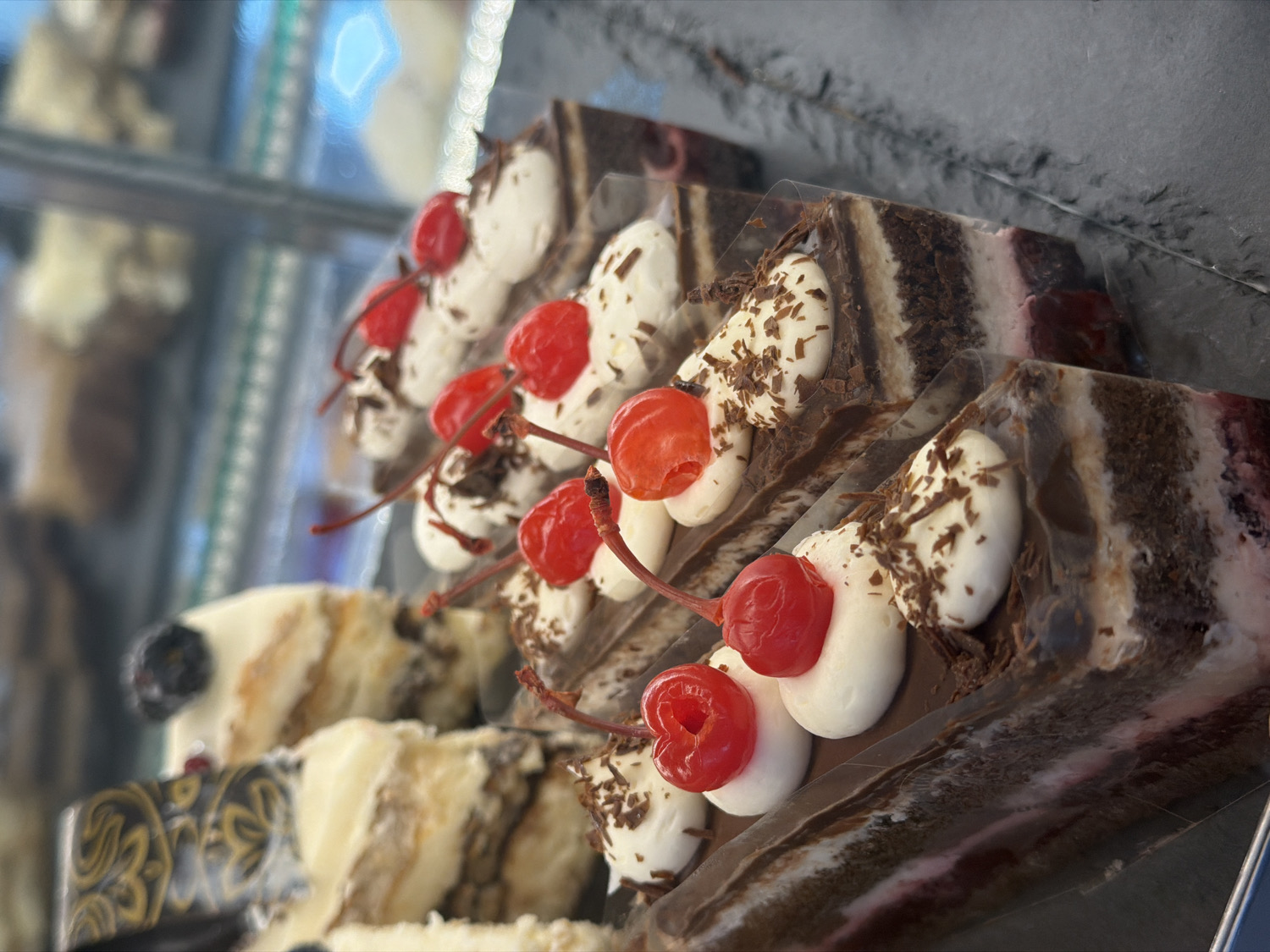 The image presents a close-up, oblique view of several individual slices of cake displayed in what appears to be a refrigerated glass case, likely in a bakery or dessert shop. The cakes are predominantly dark chocolate and cream layered desserts, reminiscent of Black Forest cake.
Each slice is topped with generous dollops of white whipped cream or frosting, sprinkled with dark chocolate shavings. A vibrant red maraschino cherry with a stem adorns each cream portion, providing a striking color contrast. The cakes exhibit distinct dark and light striped layers, suggesting rich chocolate sponge alternating with lighter cream or perhaps another cake flavor.
The slices are arranged in rows, with the ones in the foreground sharply in focus, while the background elements and some of the cakes further back are softly blurred due to a shallow depth of field. Clear plastic packaging or wrappers encase each individual slice. The cakes rest on a dark, possibly stone or metallic, surface to the right, and a decorative pattern is faintly visible on a plate or tray beneath cakes on the left side of the frame.
The lighting appears to be internal, casting soft reflections on the glass display and the glossy cherries. There are no people visible, and no specific activity other than the static display of the desserts. No discernible text is visible in the image. The time of day and weather conditions cannot be determined.