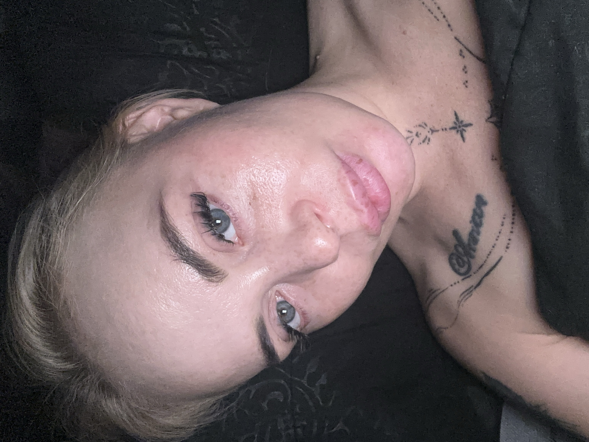 A close-up, overhead shot shows a fair-skinned woman with blonde hair and blue eyes lying down. Her face is tilted slightly upwards and to the left, and her eyes are partially closed. She has noticeable makeup, including long eyelashes, and some freckles are visible on her cheeks. The woman has tattoos on her chest and shoulder area. The background is dark and appears to be a dark fabric. The lighting is dim, and there is a slight glare on her forehead and cheeks, suggesting a flash was used.
