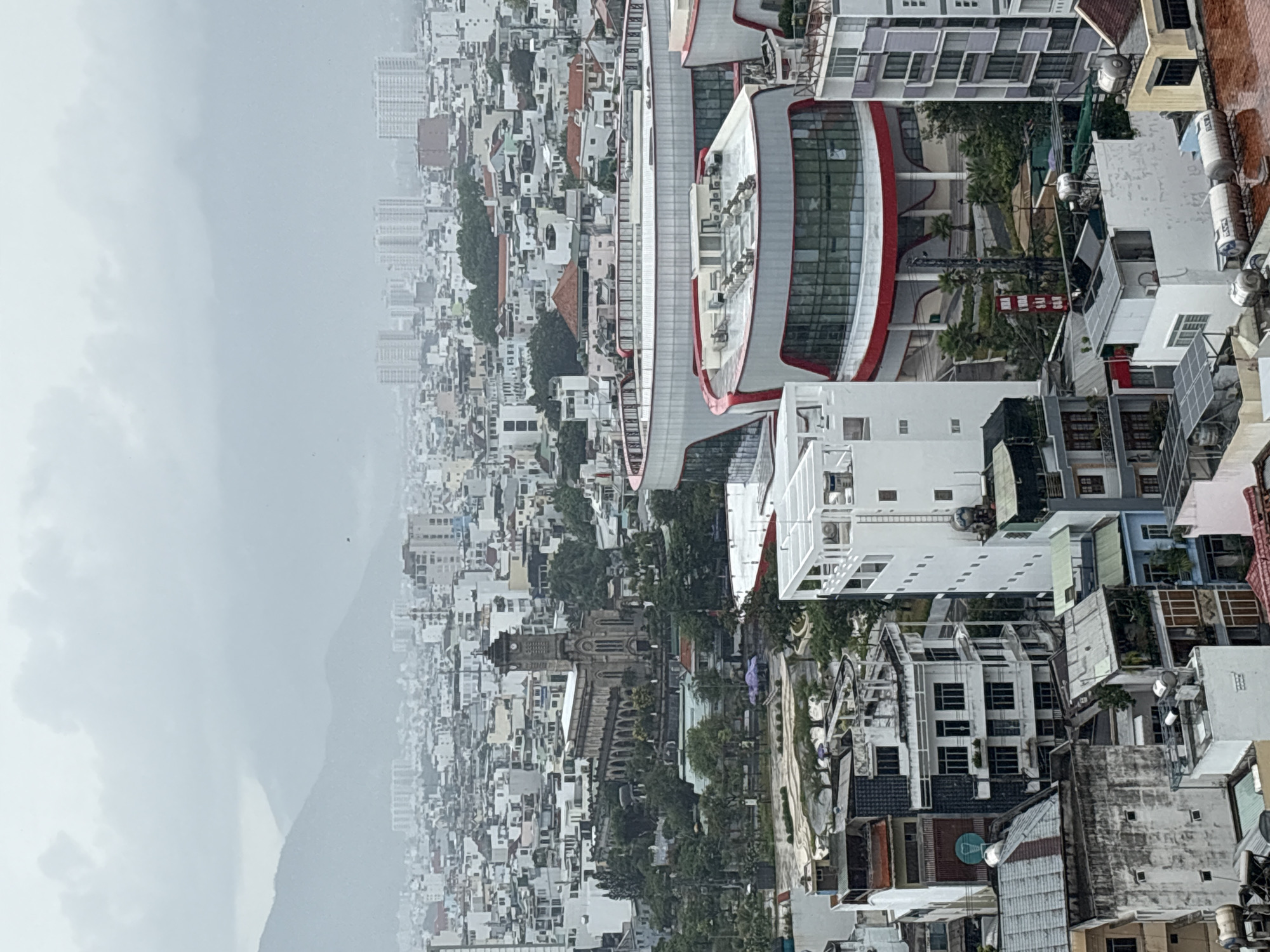 The image shows an aerial view of the city of Nha Trang Ward, Vietnam, captured on a cloudy day with a backdrop of misty mountains. The urban landscape is densely populated with buildings of varying heights and architectural styles, predominantly white and grey in color.
Notable structures visible are the Nha Trang Cathedral, characterized by its stone architecture and pointed spires, and a modern building with a curved façade and red accents. The streets are interwoven among the buildings, visible with few people or vehicles present. Trees and greenery are interspersed throughout the city, softening the urban landscape. The sky is overcast, casting a diffused light over the scene.