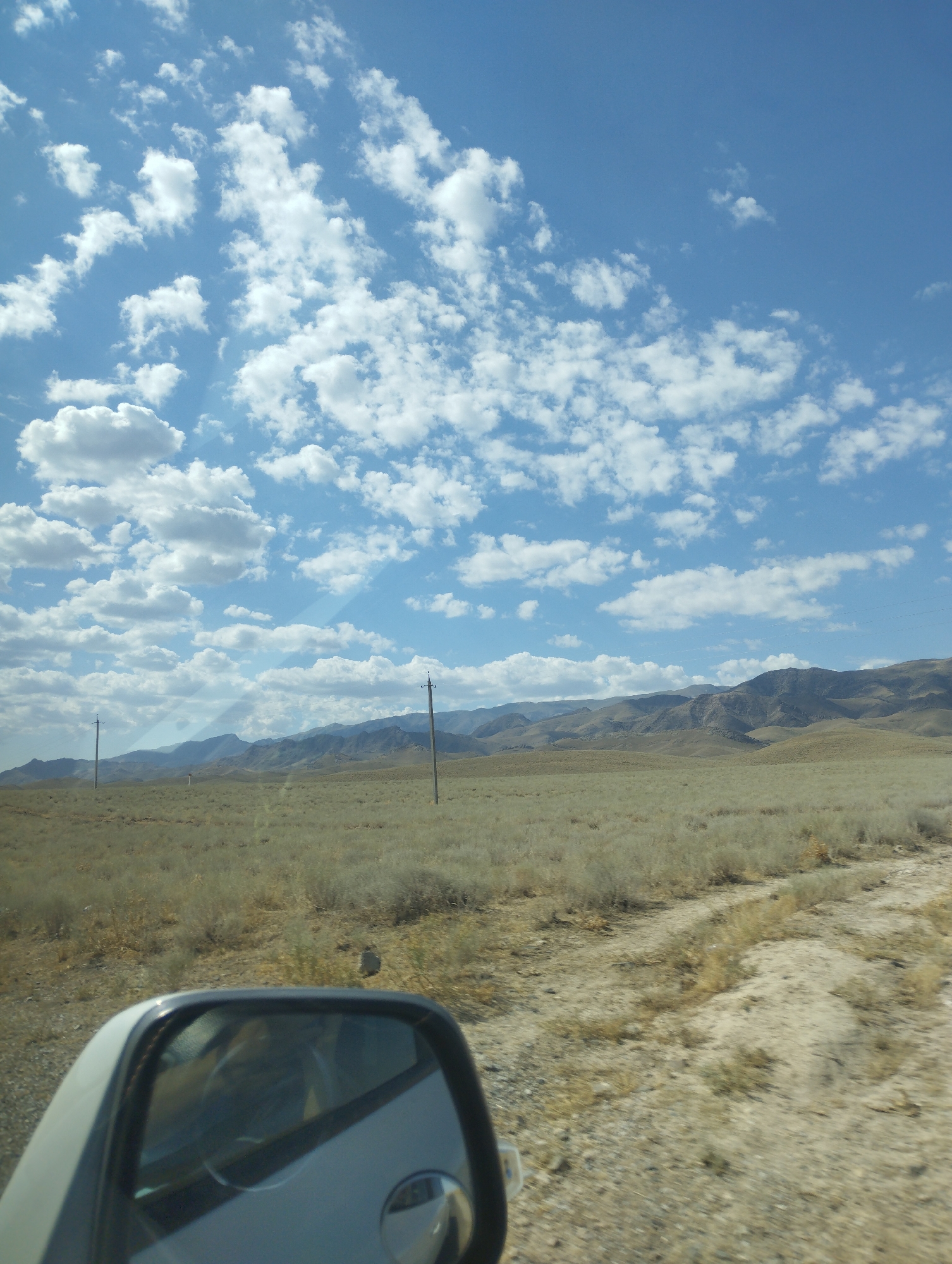 The image shows a view of a vast, arid landscape with low, rolling hills in the distance under a bright blue sky filled with fluffy white clouds. In the foreground, a dirt road or track curves through dry, sparse vegetation. A side-view mirror of a vehicle is visible in the lower left corner, reflecting part of the vehicle's interior. Two utility poles with wires are sparsely placed in the landscape, indicating some form of infrastructure. The overall scene is one of a remote, undeveloped area. The location is stated to be in the city of Temirkavuk, Uzbekistan.