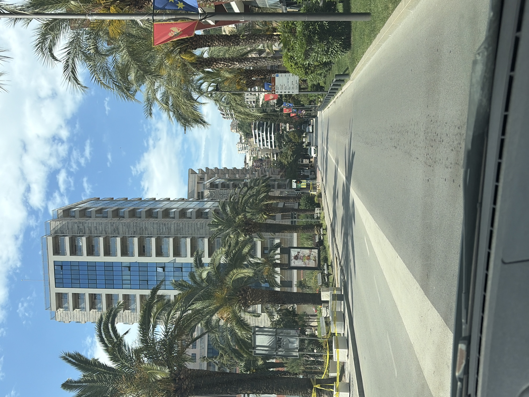 The image shows a street view in Budva, Montenegro, taken from inside a vehicle. A straight, grey asphalt road stretches into the distance, flanked by palm trees. Modern buildings line the left side of the road, including a tall structure with mirrored windows. Small white concrete barriers with yellow caution tape are along the roadside. Flags, including what appears to be the Montenegrin flag, are visible on light poles. The sky is blue with scattered white clouds.