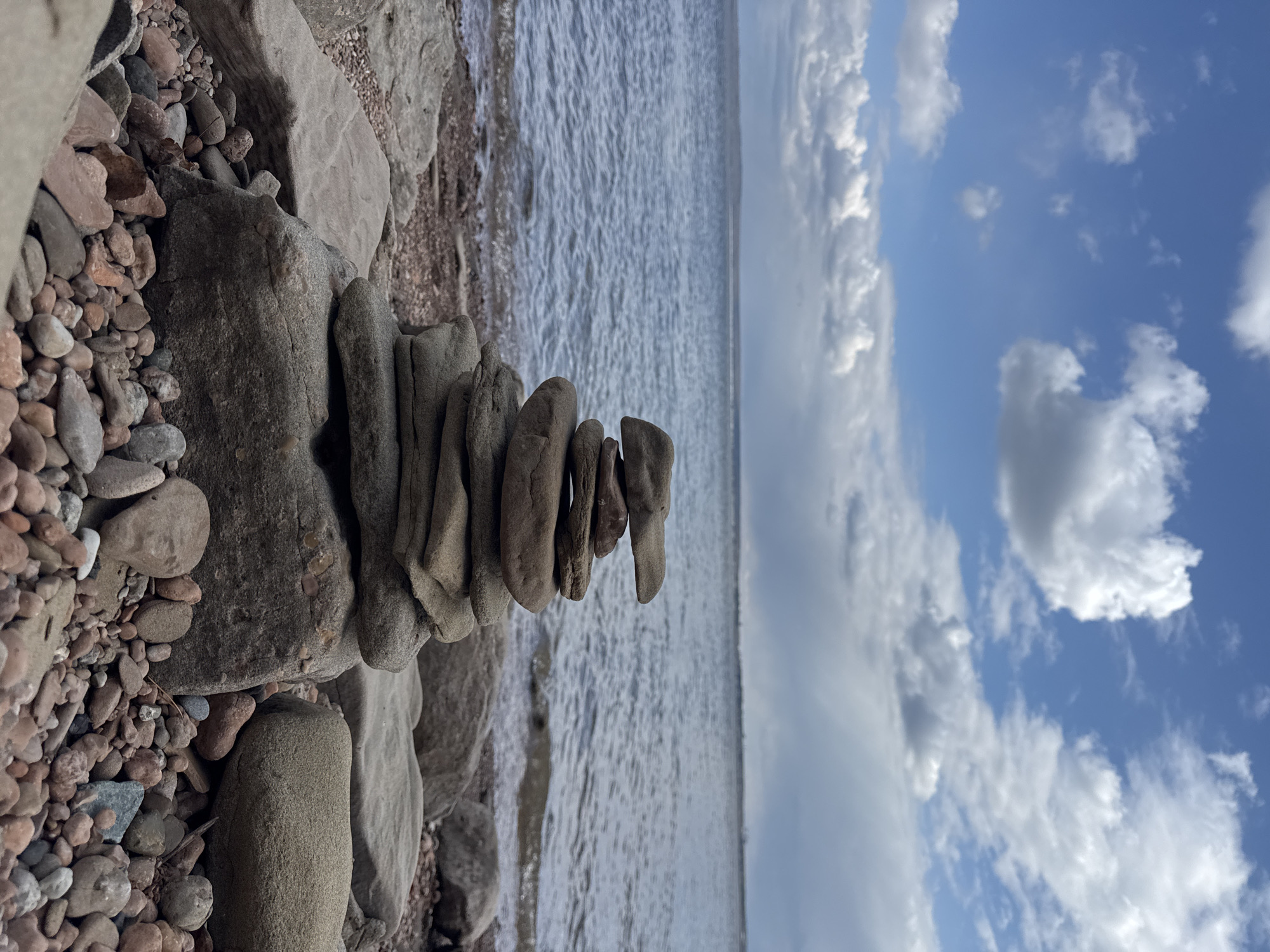 The image, when correctly oriented, depicts a serene outdoor scene featuring a rock cairn on a pebbled shore beside a large body of water under a partly cloudy sky.
In the foreground and extending along the left side, numerous small to medium-sized rocks and pebbles, displaying shades of grey, brown, and reddish-brown, form the rugged shoreline. A prominent stack of carefully balanced, smooth, greyish-brown stones, known as a cairn, stands upright in the lower-center of the frame, emerging from the rocky ground. To the right of the cairn, a vast expanse of water stretches horizontally, its surface textured with ripples that suggest gentle movement or light reflection. The horizon line is visible in the distance.
The upper portion of the image is dominated by a bright blue sky, adorned with scattered white, fluffy clouds of varying sizes. The scene is illuminated by natural daylight, indicating a daytime setting, likely late morning or early afternoon, under fair weather conditions. There are no people present, and no activities are explicitly taking place. The image conveys a sense of natural tranquility and balance. No visible text is present within the frame. The location is a natural, outdoor environment, consistent with a lake or sea shore in Noviy, Russia.