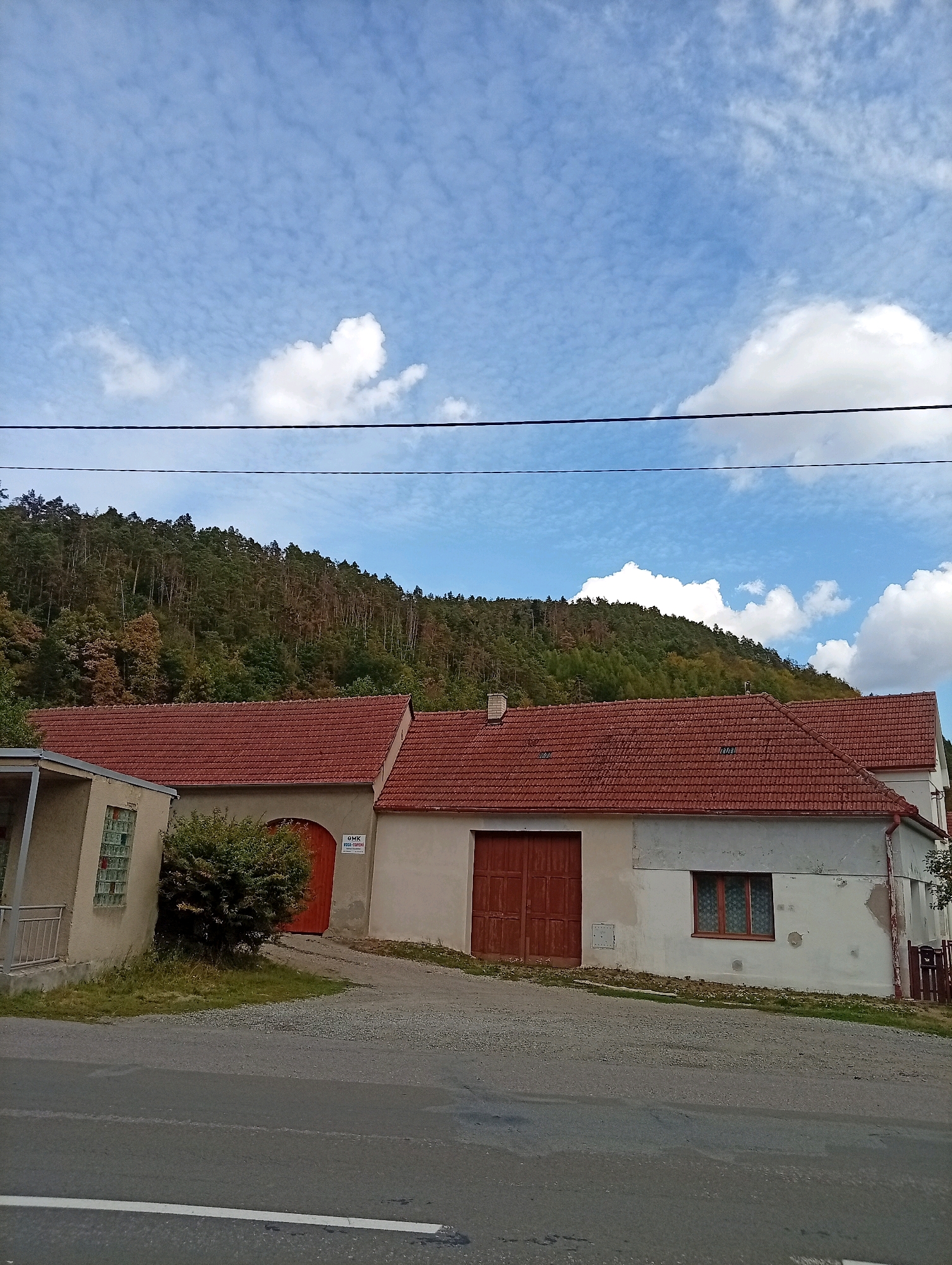 The image captures a street view in Borač, Czechia, on a partially cloudy day. In the foreground, a paved road runs horizontally across the bottom of the frame, with white lines indicating lanes. Behind the road, a gravel driveway leads up to a collection of buildings with red tile roofs.
To the left, a smaller, lighter-colored building with a covered entrance and a fence is visible. Adjacent to it, a larger building features a large, arched wooden door. Further to the right, another building, primarily white, has a smaller wooden door and a window with decorative curtains. The architecture suggests an older, possibly rural or semi-rural setting.
The background is dominated by a densely forested hillside, with a mix of deciduous and coniferous trees displaying varying shades of green and some autumnal colors. The sky above is a light blue, interspersed with scattered white clouds, some of which are puffy and distinct, while others form wispy streaks. Overhead power lines are visible against the sky, stretching horizontally across the upper portion of the image. No people are present in the scene.