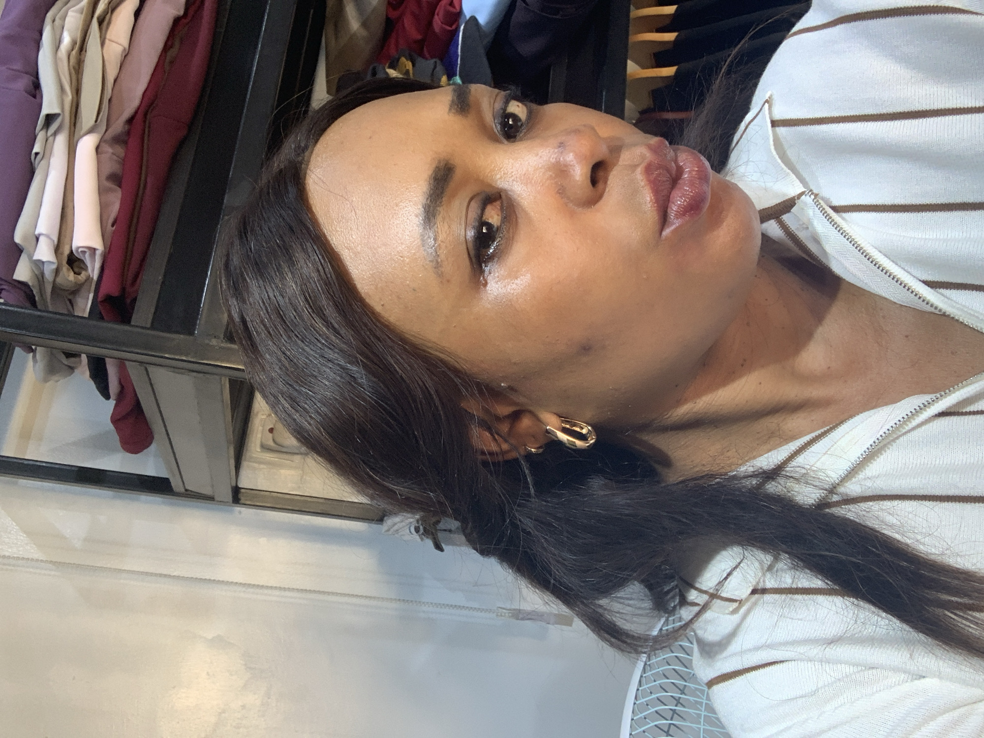 A woman with dark hair, wearing a striped top and gold earrings, is positioned in front of a clothing rack, possibly in Narayi, Nigeria. The woman is looking at the camera and pouting. There are no visible landmarks or environmental details to definitively place her in Narayi.