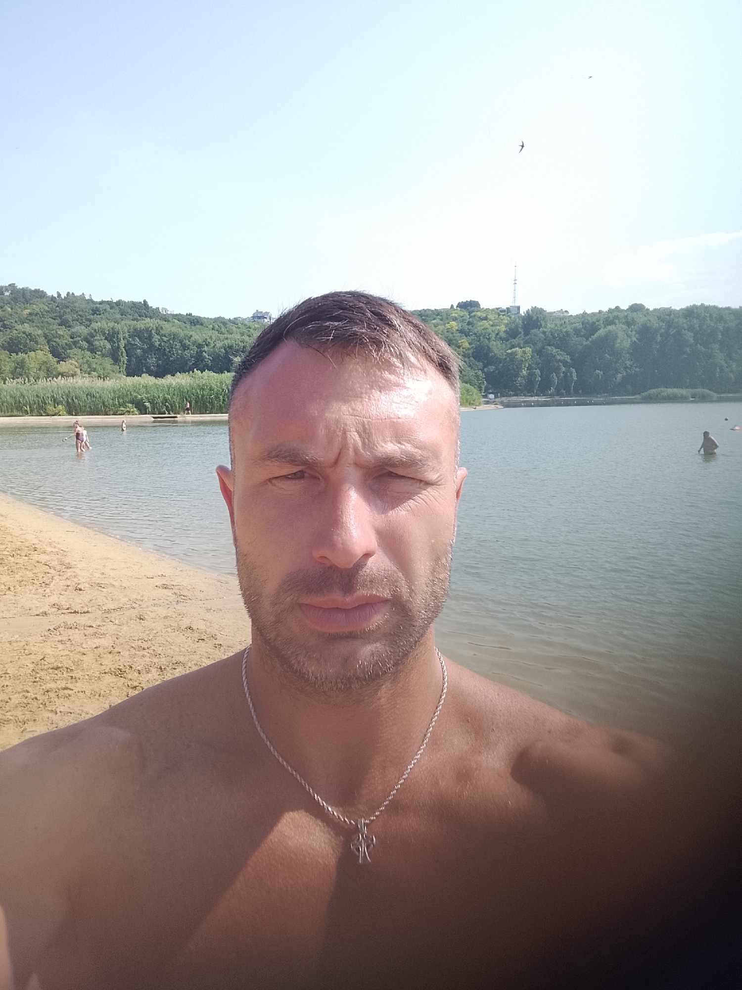 A man is taking a selfie at the beach. He is shirtless and wearing a silver necklace with a cross pendant. In the background, there is a body of water with other people swimming or wading. The beach is sandy, and behind the water, there is a wooded area. The sky is clear and blue. The location is Lake Valea Morilor in Chișinău, Moldova.