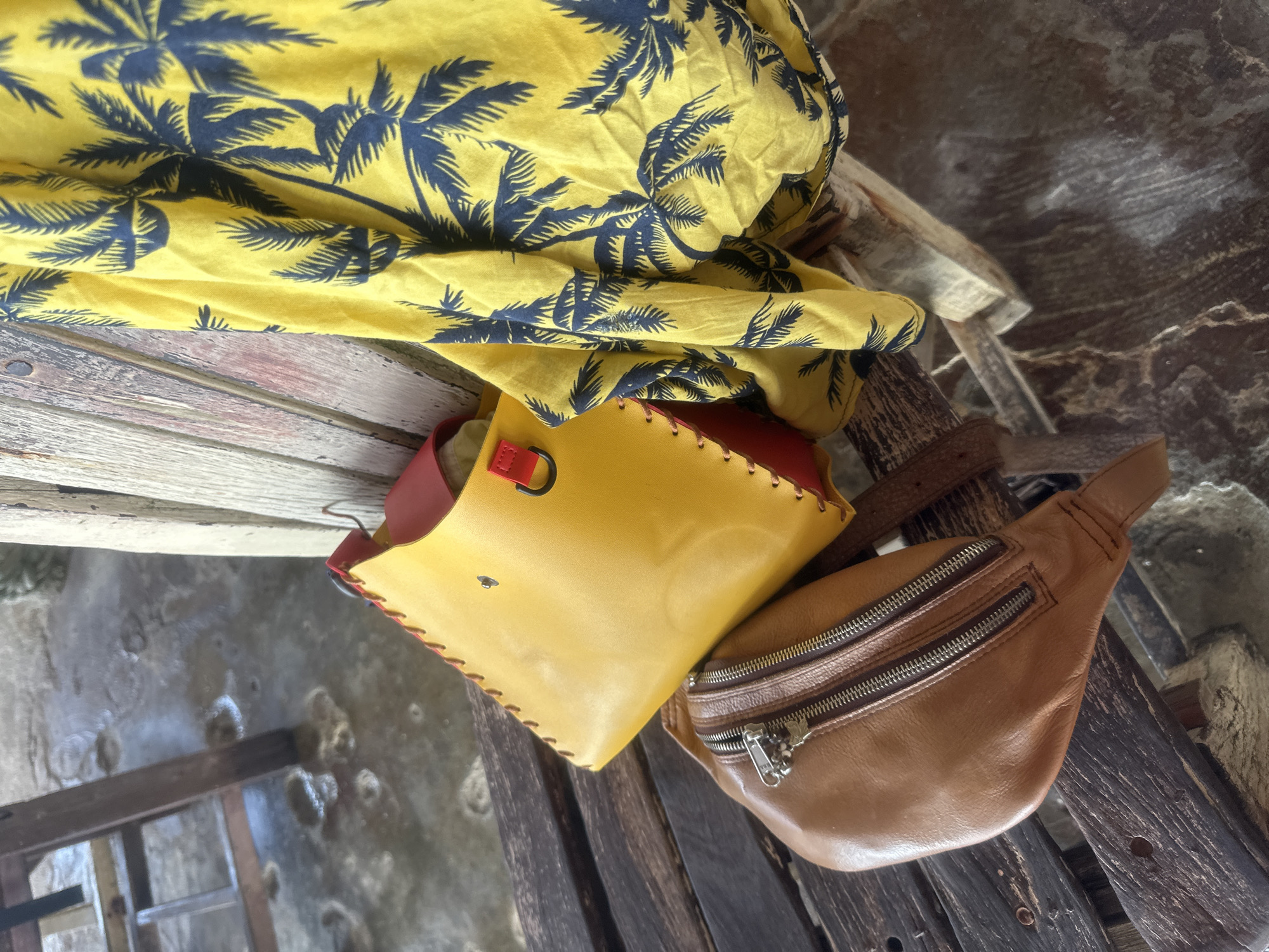 A yellow leather purse with red accents and a brown leather fanny pack with double zippers rest on a wooden bench, alongside a yellow shirt with a dark blue palm tree print. The bench is situated on a weathered wooden surface, possibly outdoors, with glimpses of a sandy or concrete ground speckled with water and debris. In the background, a wooden chair leg is visible. The scene appears to be in a relaxed, possibly tropical environment, consistent with the location of ตำบลเกาะพะงัน, Thailand, known for its beaches and relaxed atmosphere.