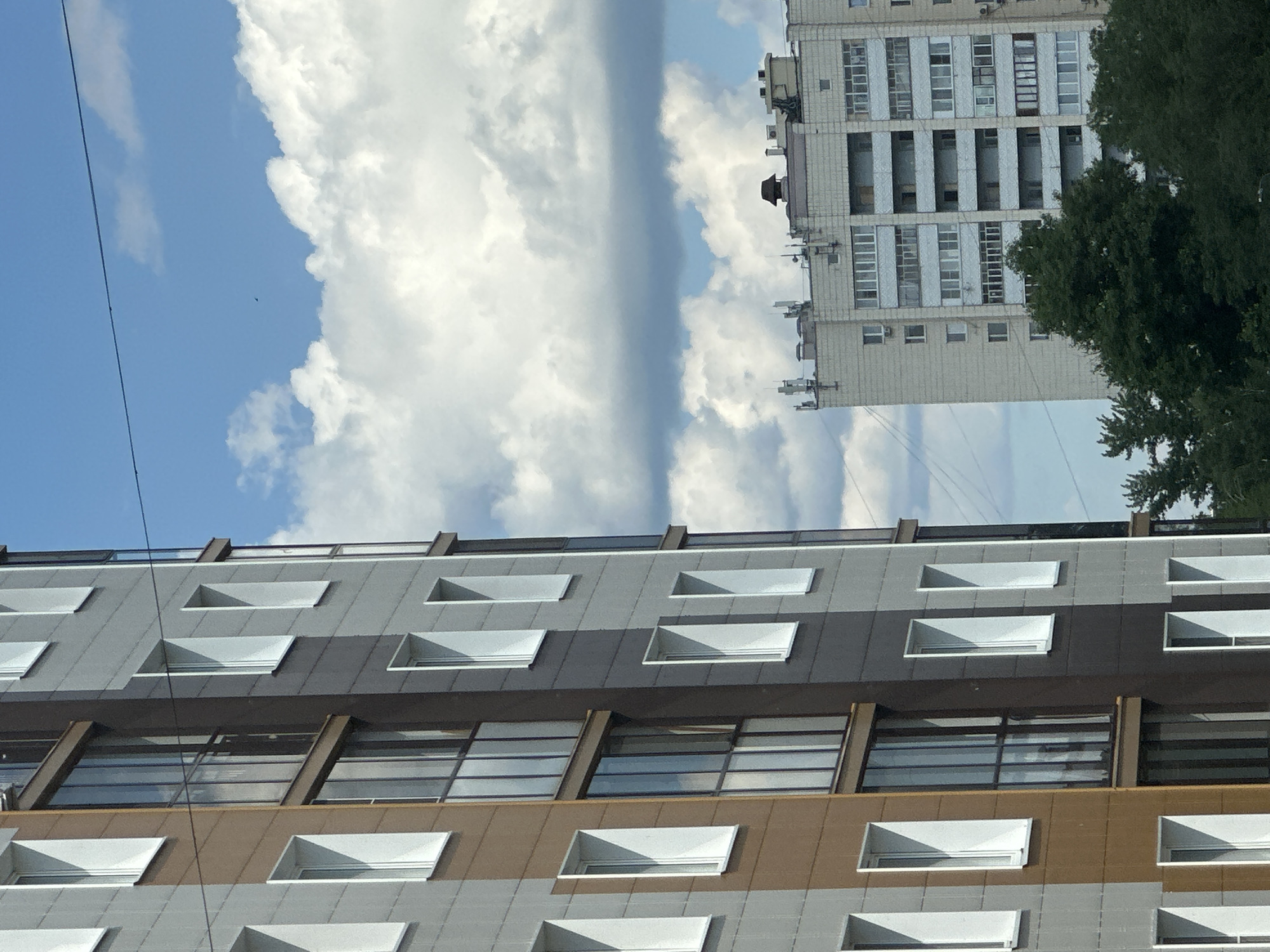 The image shows a close-up view of a modern apartment building in Kazan, Russia, on a partly cloudy day. The building on the left has a facade with alternating panels of light gray and brown, punctuated by numerous white rectangular windows and some glassed-in balconies. The building on the right, further in the background, is a lighter-colored, older style apartment block with many windows and some visible antennae on its roof. Lush green trees are visible in the foreground and at the base of the background building. The sky is a vibrant blue with large, fluffy white clouds. A thin black wire stretches diagonally across the upper portion of the image. There are no people visible in the frame.