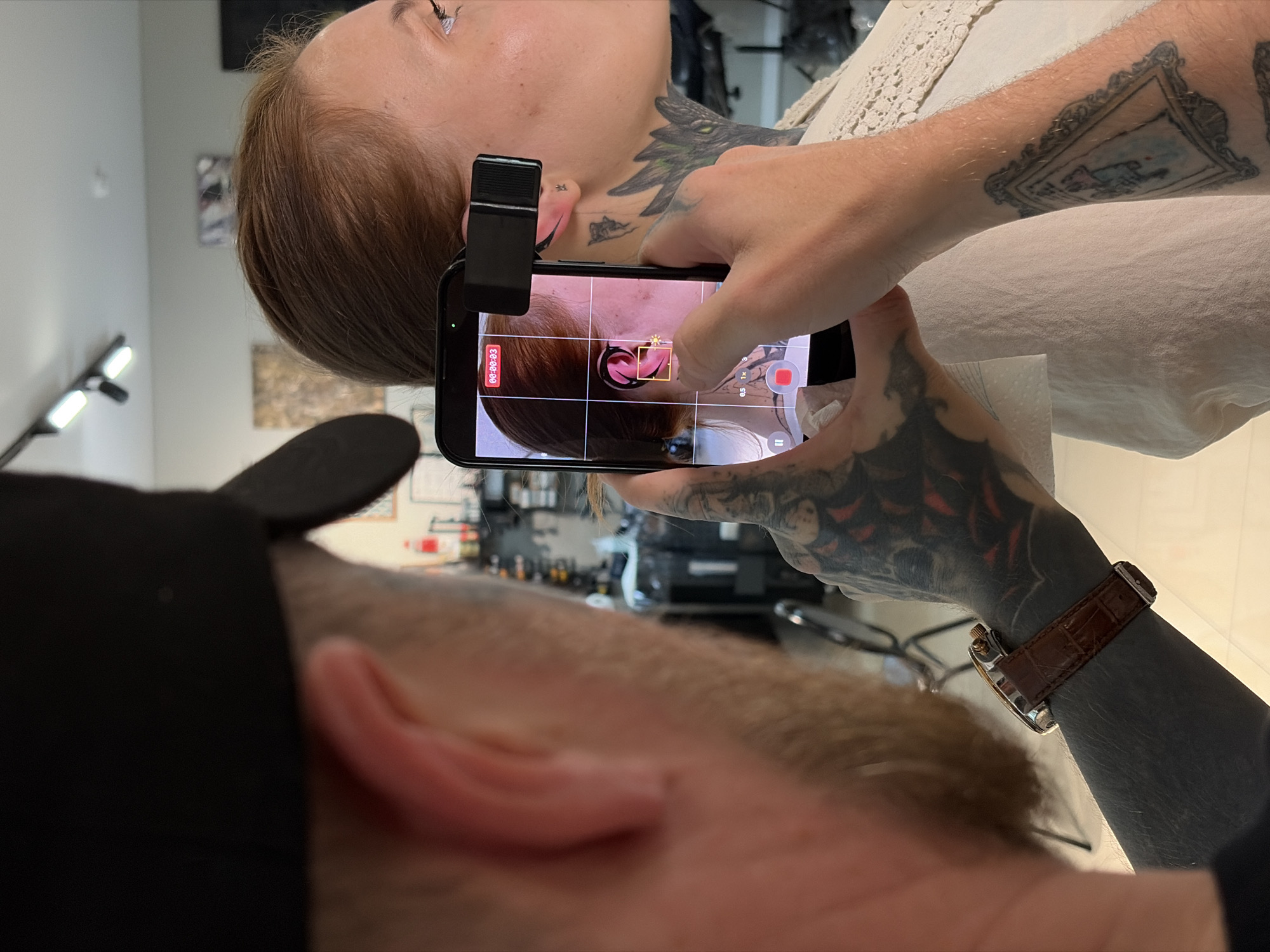 A person wearing a black baseball cap is holding a smartphone to record a woman who has a small stud earring in her left ear. The smartphone screen shows a close-up of the woman's ear with a yellow square marking a target area. The woman has a tattoo on her neck and upper chest. The background appears to be an indoor commercial space, possibly a tattoo or piercing studio, with shelves of items visible. The lighting suggests an artificial indoor environment. There is no specific information in the image to indicate the location is Izhevsk, Russia.