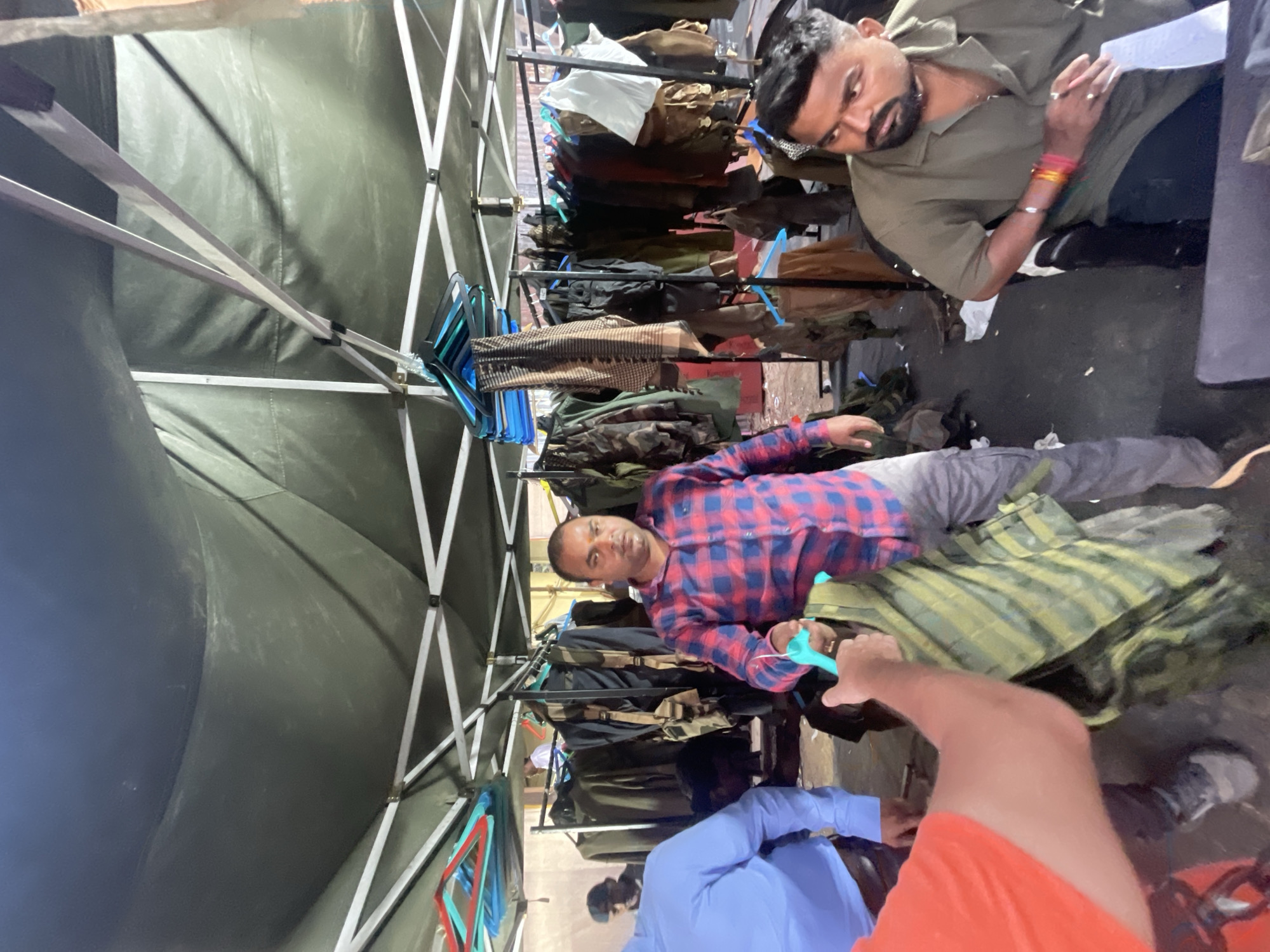 The image shows an indoor scene in what appears to be a makeshift tent or pop-up structure. Several men are present. One man is lying down, wearing a red and blue plaid shirt and grey pants. Another man is kneeling and appears to be examining paperwork or clothing; he is wearing a dark green shirt and has wristbands. Two more men are partially visible near the bottom of the frame, wearing light blue and orange shirts respectively.
A rack filled with various articles of clothing, possibly jackets or uniforms, runs along the center of the frame, creating a makeshift shop or storage area. The structure around the men is supported by metal poles and covered in green fabric. The floor appears to be concrete.
The specific location in Mira-Bhayander, India, cannot be determined from the image alone.