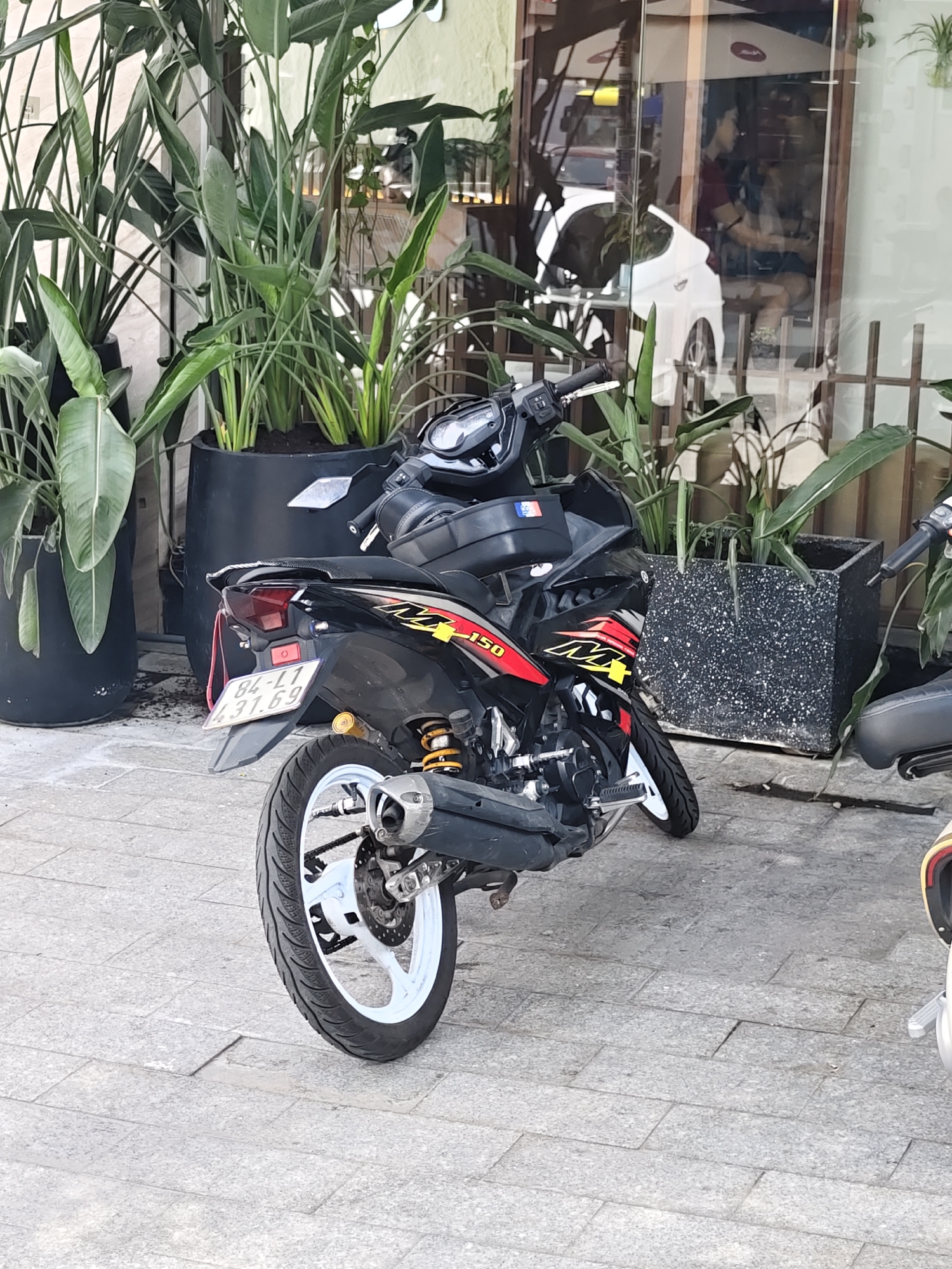 In Nha Trang Ward, Vietnam, a black and red motorcycle is parked on a paved outdoor area. The motorcycle has a license plate that reads "84-L1 431.69". It is positioned near large potted plants with long, green leaves and a building with a reflective glass facade. Through the glass, several people are visible inside, and a white car is parked. The overall environment suggests a commercial or residential area with greenery.