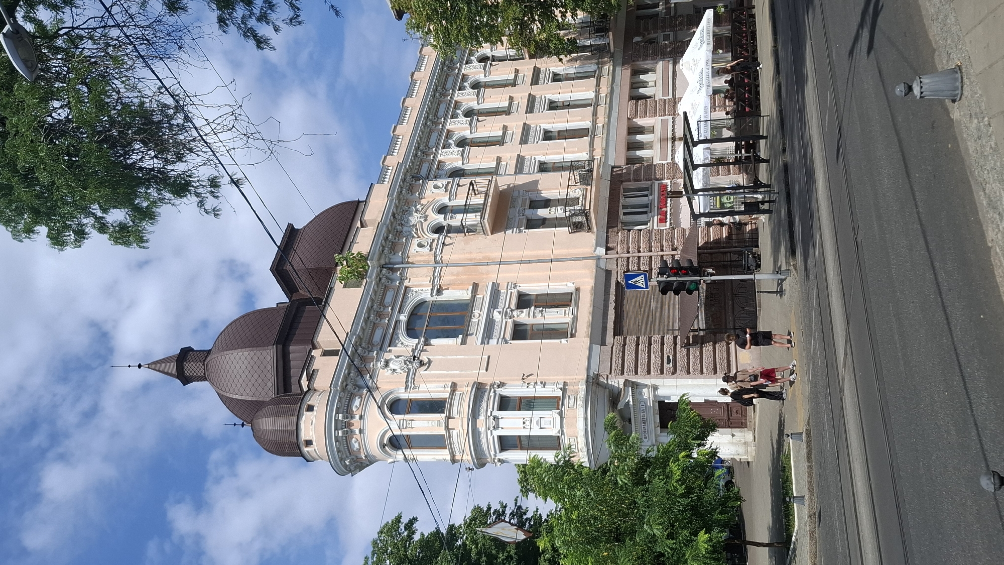 An eye-level, medium shot captures a light pink, ornate building with a brown domed roof in an urban setting under a blue sky with scattered white clouds. The building features multiple windows, balconies with decorative railings, and intricate architectural details along its facade. In the foreground, a street with tram tracks is visible, along with a sidewalk and a pedestrian crossing with a traffic light showing red. Three individuals stand on the sidewalk near the crossing. To the right of the building, an outdoor seating area with umbrellas and tables suggests a cafe or restaurant. Trees line the sidewalk, and power lines crisscross overhead. The lower portion of the building has a textured stone base.
