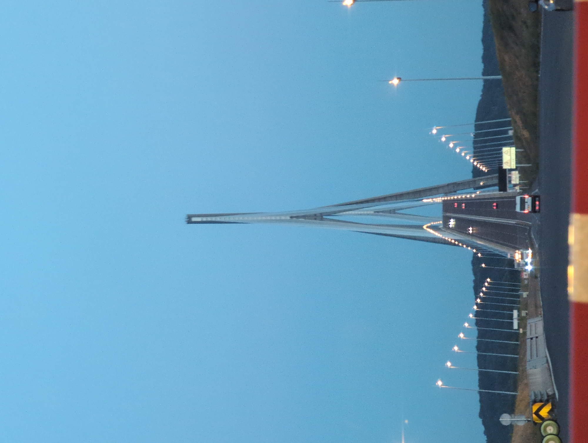 The image captures a frontal view of the dramatic, modern Pont de Normandie cable-stayed bridge at dusk, located in Sandouville, France. The sky is a clear, pale blue, transitioning from day to night. The bridge's central pylon, a towering, slender structure with a distinctive A-frame design, dominates the center of the frame, tapering upwards and against the sky. Cables extend from the pylon, supporting the roadway.
The roadway of the bridge is visible, with vehicles appearing as streaks of light due to the long exposure. Several vehicles, including a white van and what appear to be cars, are moving across the bridge. Rows of bright streetlights line both sides of the roadway, creating a luminous path. These lights are also reflected on the wet surface of the road.
On the left side of the image, a concrete barrier separates the roadway from the embankment. Yellow and black chevron signs and circular white signs are mounted on poles along the embankment. On the right, a sloping grassy hillside is visible, also lined with streetlights. The overall environment suggests a coastal or riverine setting, characteristic of the bridge's location, with a dark, wooded area forming the horizon behind the lights. In the foreground, a red and white barrier or railing is partially visible, indicating the vantage point of the photographer.