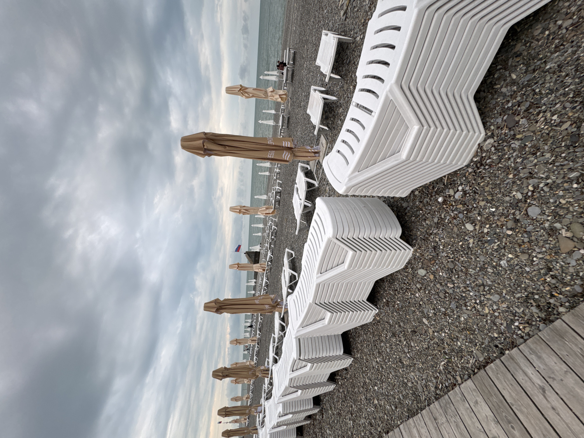 The image depicts a pebbled beach in Sochi, Russia, under a cloudy sky. Rows of closed, tan beach umbrellas and stacked white plastic beach chairs are lined up on the beach. A wooden boardwalk runs along the right side of the frame. In the distance, people can be seen on the beach, and a Russian flag is visible. The overall scene suggests an off-season or quiet day at the beach.