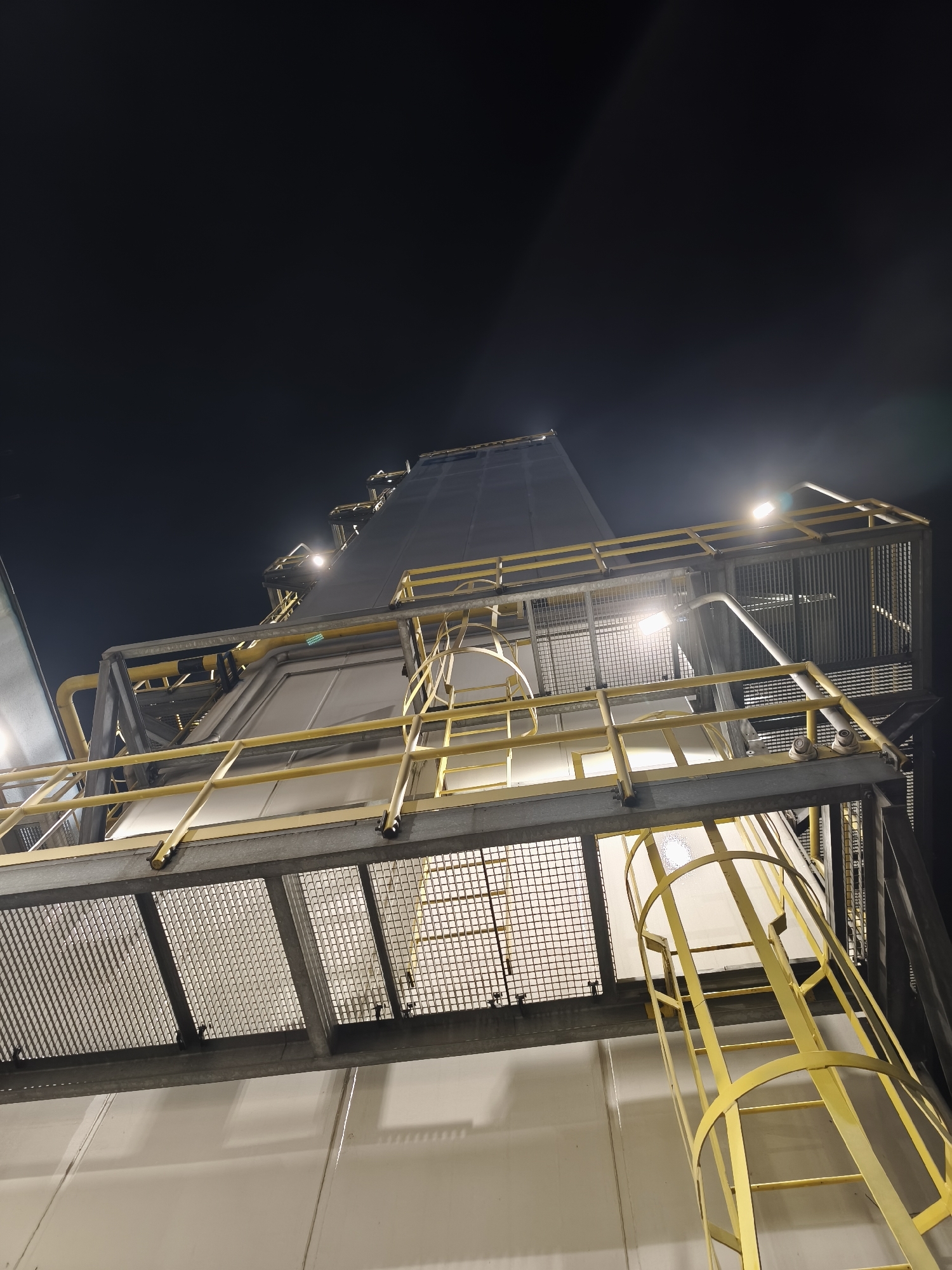 The image depicts a large, multi-level industrial facility at night, observed from a low-angle perspective looking upwards. The primary structure is composed of vertical, light-colored (off-white/pale grey) paneled walls, suggesting a building, silo, or processing tower. Multiple platforms and walkways, constructed from grey metal grating and structural beams, are integrated into the building's exterior at various elevations.
Prominent yellow safety railings, consisting of horizontal bars, are installed along the perimeter of these platforms and stairways. A yellow caged ladder, designed for safe vertical ascent, is visible on the lower right, extending upwards along the facility's side. Yellow pipes run horizontally and vertically along sections of the structure, indicating functional utility lines.
The scene is illuminated by several bright, artificial floodlights mounted on the platforms, casting intense light upwards onto the facility and against the dark night sky. Some lens flare is evident from these light sources. No individuals are present within the frame. The overall impression is one of an operational industrial site after dark, located in the city of Unknown, Unknown.