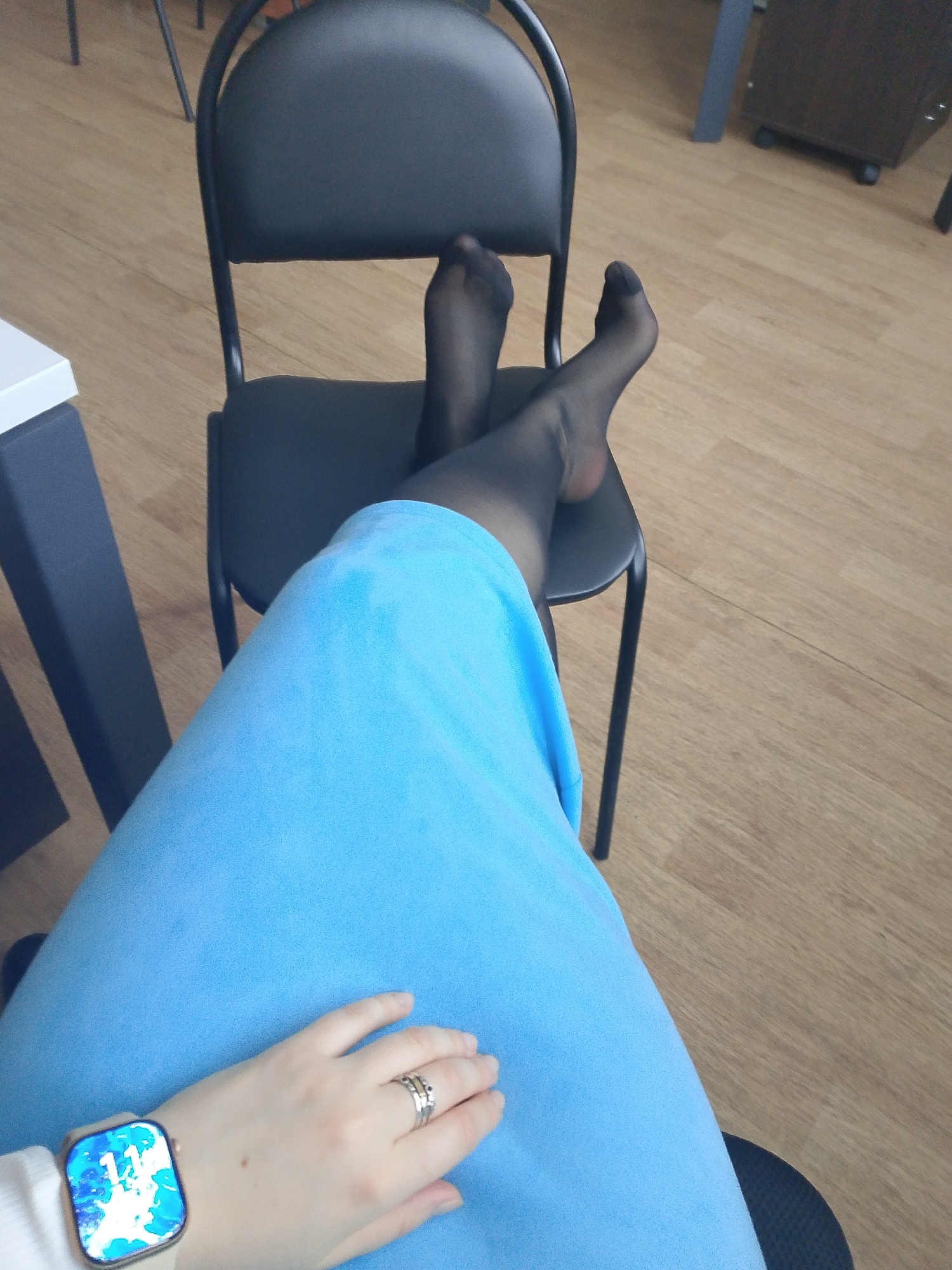 A person wearing a light blue dress and black stockings is sitting in an office chair. Their feet, covered in stockings, are resting on another chair. The person is wearing a smartwatch and rings on their left hand. The location appears to be an office setting with a wooden floor. There is no evidence in the image to pinpoint this location in the city of Izhevsk, Russia.