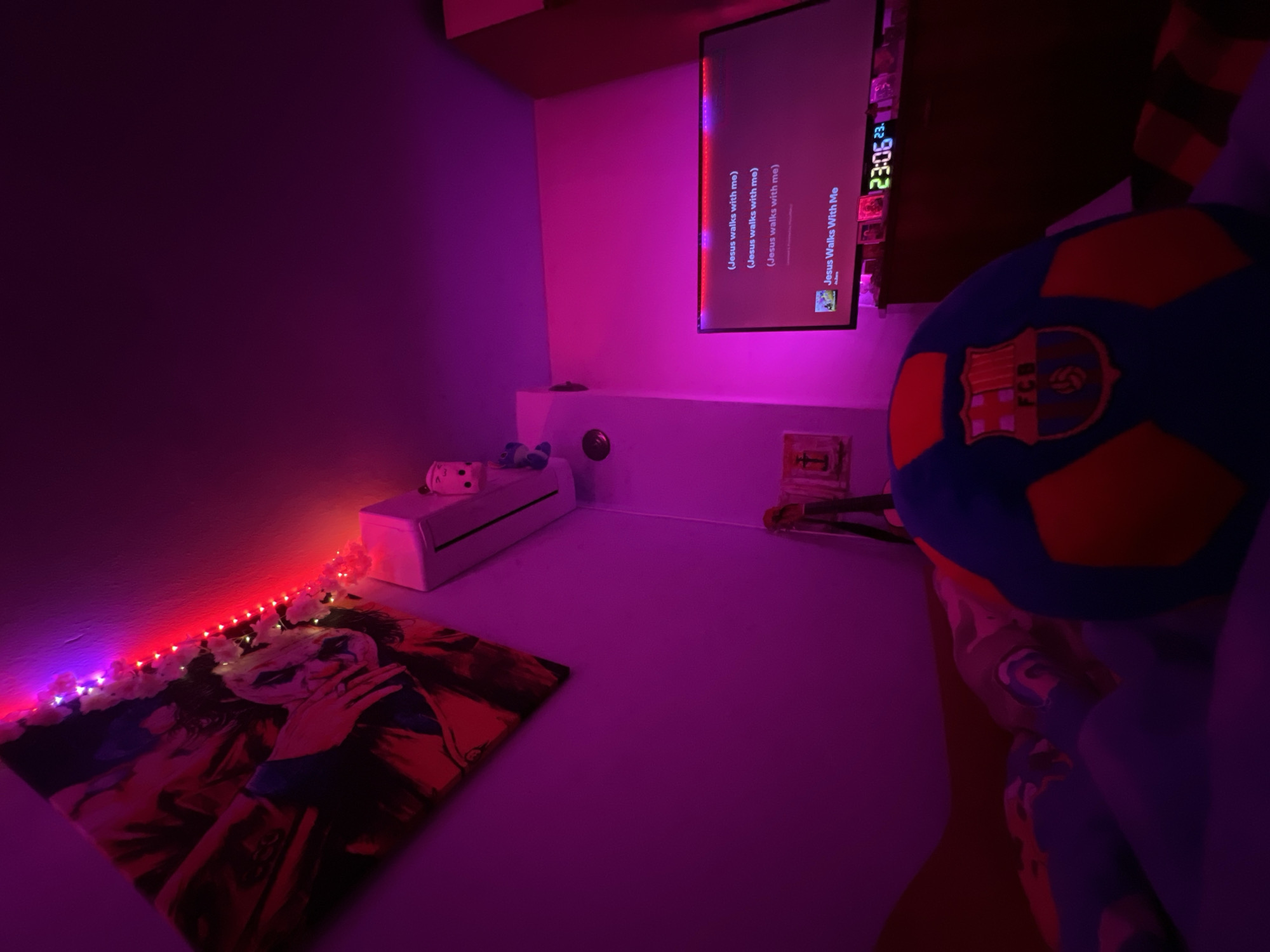 The scene is an interior residential room, presumably located in Debelt, Bulgaria, illuminated primarily by strong magenta, purple, and red ambient lighting. The perspective is low, suggesting the camera is placed on a bed or floor, looking upwards and across the room.
On the left wall, a large, rectangular framed artwork is mounted at an angle, depicting Joaquin Phoenix as the Joker from the 2019 film. The painting's colors are heavily influenced by the room's lighting, appearing in shades of red, orange, and dark blue. Along the intersection of the wall and ceiling, above the painting, runs a strip of LED string lights, predominantly red, with a segment shifting to purple/blue. Small white decorative elements, possibly artificial flowers or leaves, are intertwined with sections of the lights.
Below the lights and to the right of the painting, a white, wall-mounted air conditioning unit is visible. On top of the unit, two small, white, stylized figures with black facial features are perched. Further down the wall, below the AC unit, a small, dark, circular object (possibly a sensor or vent) is present, and below that, a small religious icon resembling a cross is affixed to the wall.
Towards the right side of the room, a flat-screen television is displaying song lyrics. The screen prominently shows "(Jesus walks with me)" repeated three times, followed by the song title "Jesus Walks With Me" and the artist "A_ _ _ _ _" (partially obscured). Below the television, on a dark-colored piece of furniture (likely a dresser or TV stand), a green digital clock displays "23:06" (11:06 PM), with an additional "23." in red to its right. Several other small, dark objects, possibly framed photos or decorative items, are also arranged on the furniture.
In the immediate foreground, filling the lower portion of the frame, is a blue and orange soccer ball with the FC Barcelona (FCB) crest clearly visible. The ball is slightly out of focus. Remnants of bedding or a blanket are visible beneath and behind the soccer ball, featuring dark and light striped or patterned fabric. No people are visible in the image.