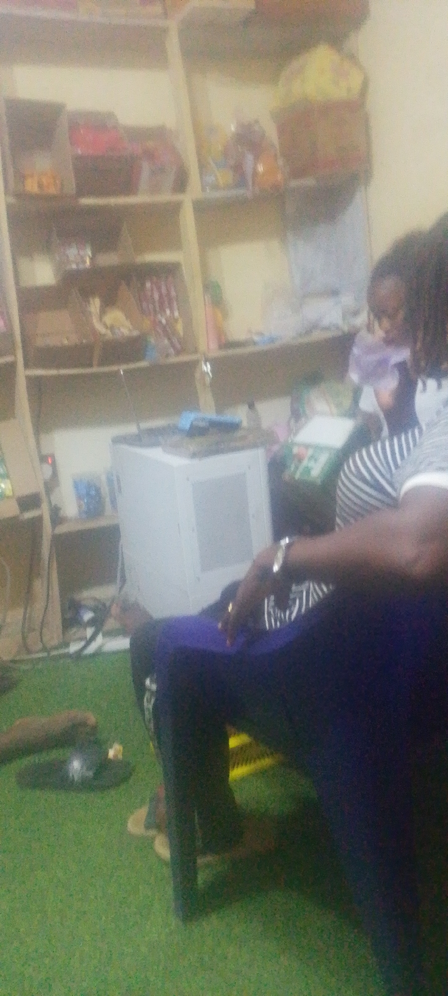 An interior view reveals a person seated in what appears to be a room in Adankolo, Nigeria. The setting is a room with a green floor, suggestive of artificial turf or carpet. A tall, light-colored shelving unit occupies the background, displaying various items including baskets and packages of goods. A white appliance is situated on the floor near the shelving unit. The seated individual is wearing a striped shirt, dark pants, and a watch on their wrist. The image clarity is poor, making it difficult to discern finer details. The angle and composition suggest a candid, possibly hastily captured, shot.