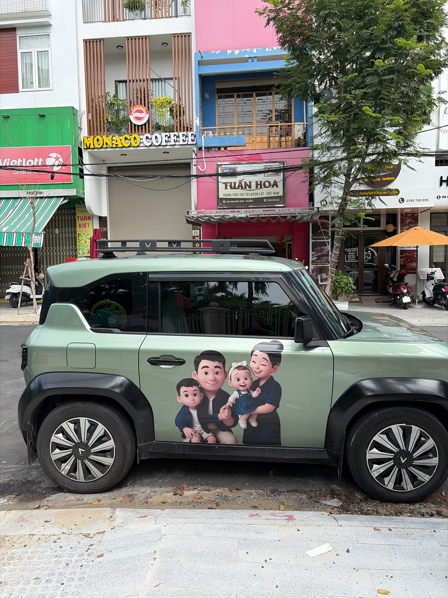 The image shows a light green electric vehicle parked on a street in Đà Nẵng, Vietnam. The vehicle has a cartoon sticker of a family of four on its side. In the background, there are several buildings with various businesses, including "Monaco Coffee," "Vietlott," and "Tuan Hoa" which appears to be a gallery. The street is paved with concrete and cobblestones, and there are trees and a bicycle on the sidewalk. The scene appears to be a typical street view in a Vietnamese city.