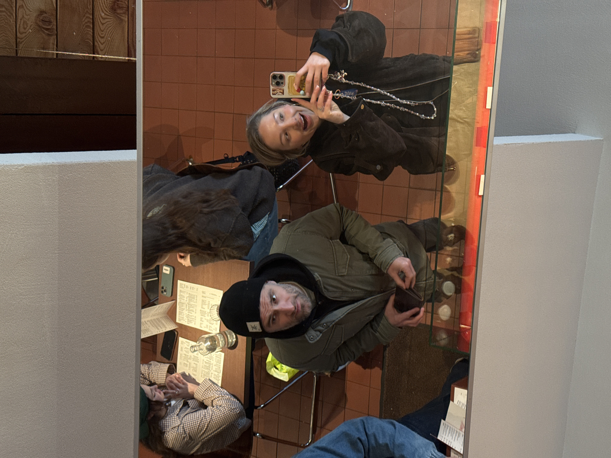 The image captures an indoor scene within what appears to be a cafe or restaurant in Moscow, Russia. The perspective is unusual, suggesting the photo was taken in a mirror or reflective surface, causing a slight distortion and reversal of the scene.
Two main individuals are visible. On the left, a young woman with blonde hair is taking a selfie with her smartphone. She has a playful expression, smiling broadly with her tongue slightly out, and is wearing a dark jacket and a decorative necklace with a visible "DUCH" charm. Her phone case is adorned with stickers.
Seated opposite her is a man with a beard, wearing a dark beanie with a small white logo, a black hoodie, and a military-green jacket. He looks directly at the camera with a curious or slightly surprised expression, holding a dark brown wallet open with both hands.
They are seated at a dark wooden table, which holds several white paper menus (featuring Cyrillic script, though unreadable) and a clear glass bottle. The floor is tiled in a reddish-brown pattern. In the background, wooden wall panels are visible on the far left, and plain white/grey walls on the right. A glass partition or display with a red frame and white rectangular elements runs vertically on the right side of the image, reflecting parts of the interior.
Partial views of other individuals include an arm and torso in a checkered shirt on the far left, and legs in blue jeans at the bottom right. The lighting is warm, suggesting an indoor evening setting. The activity appears to be a social gathering, possibly dining, where one person is spontaneously taking a photo.